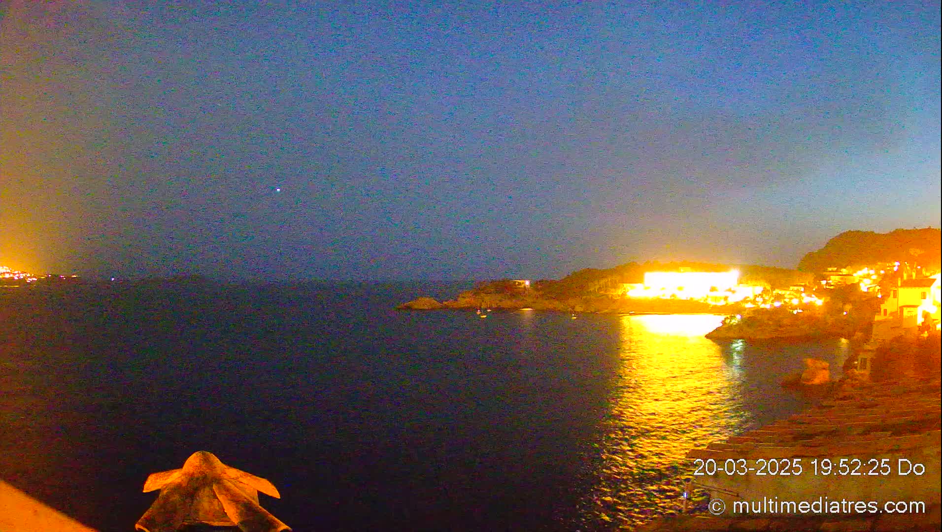 A nighttime view shows a dark body of water reflecting the lights of a coastal town under a twilight sky.