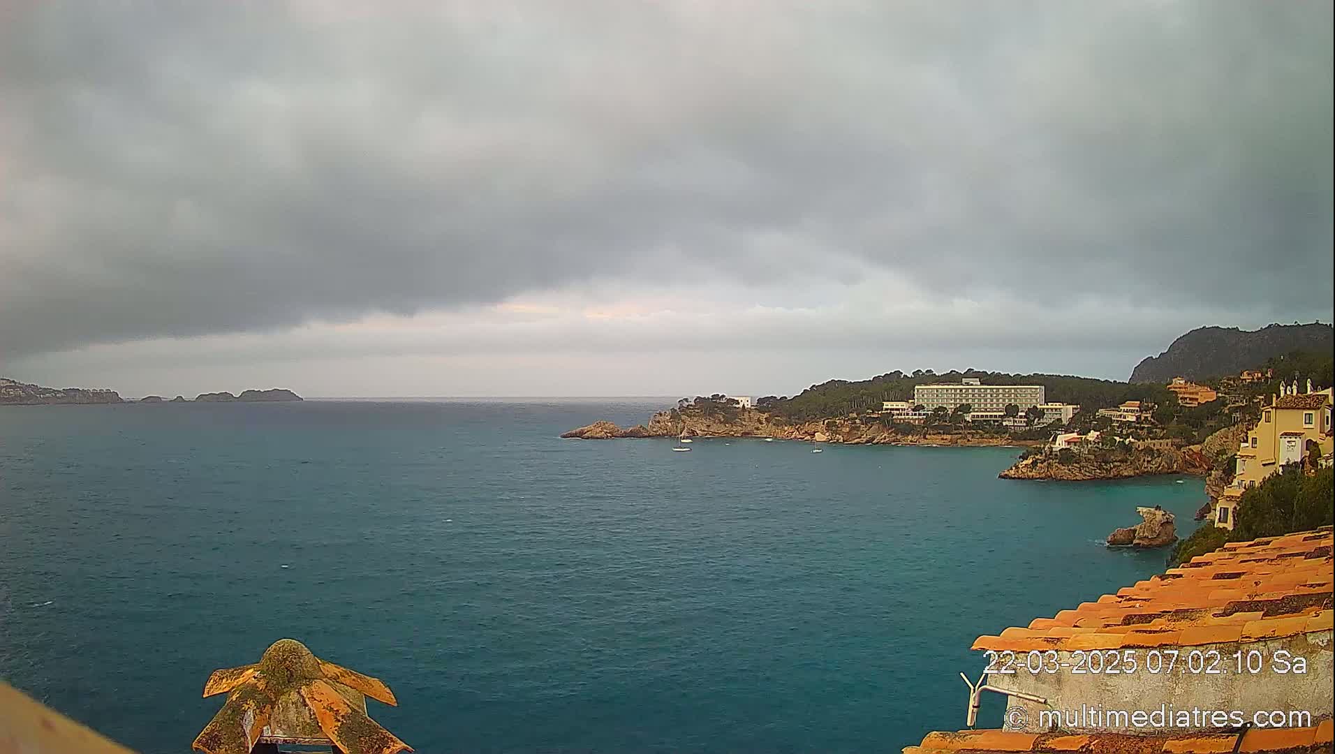 A coastal scene under an overcast sky shows a body of blue water with a rocky shoreline featuring buildings and a few small boats.