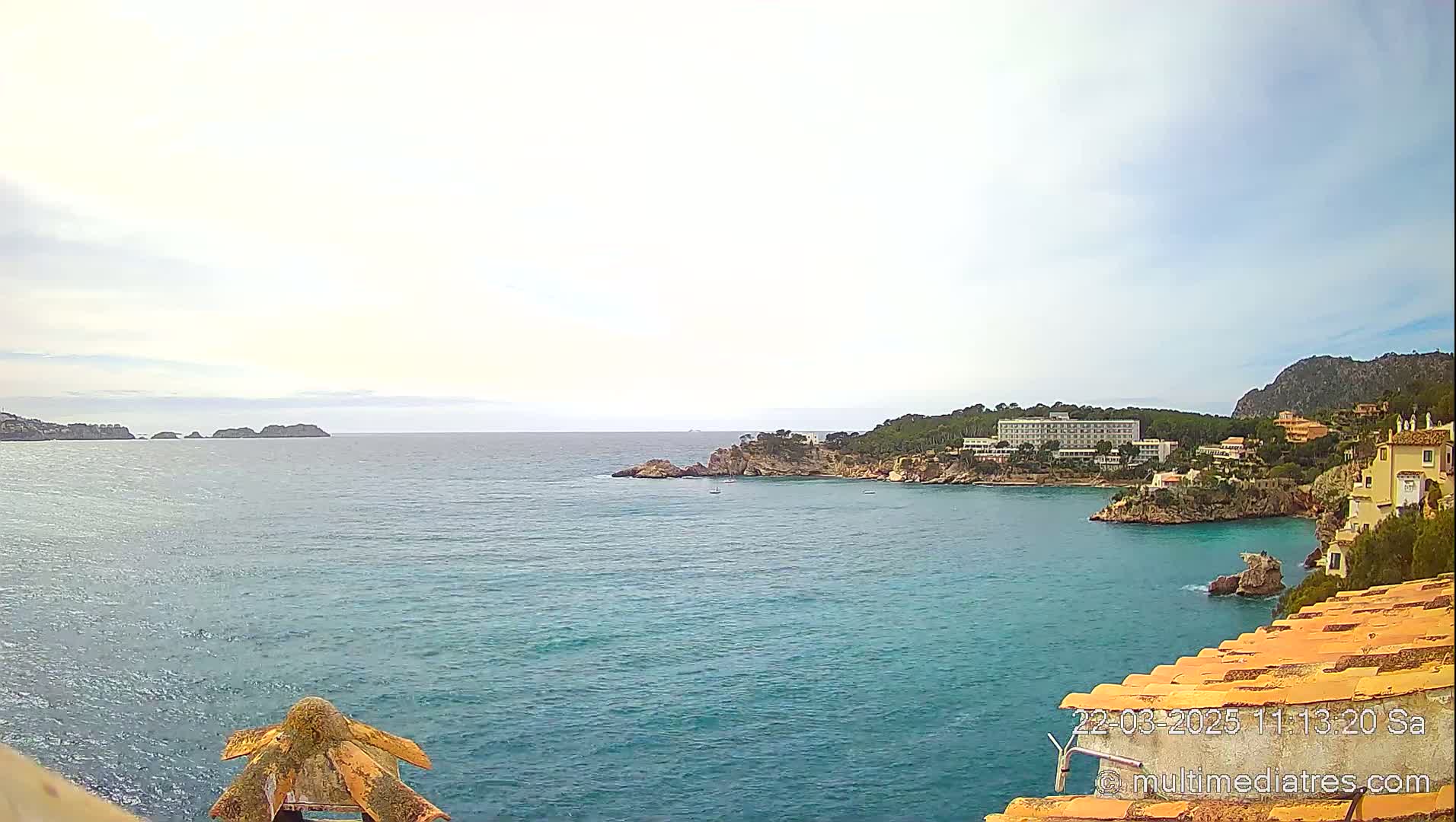 A tranquil coastal scene under a partly cloudy sky shows a calm turquoise sea with rocky inlets, distant islands, and buildings nestled along a hillside.