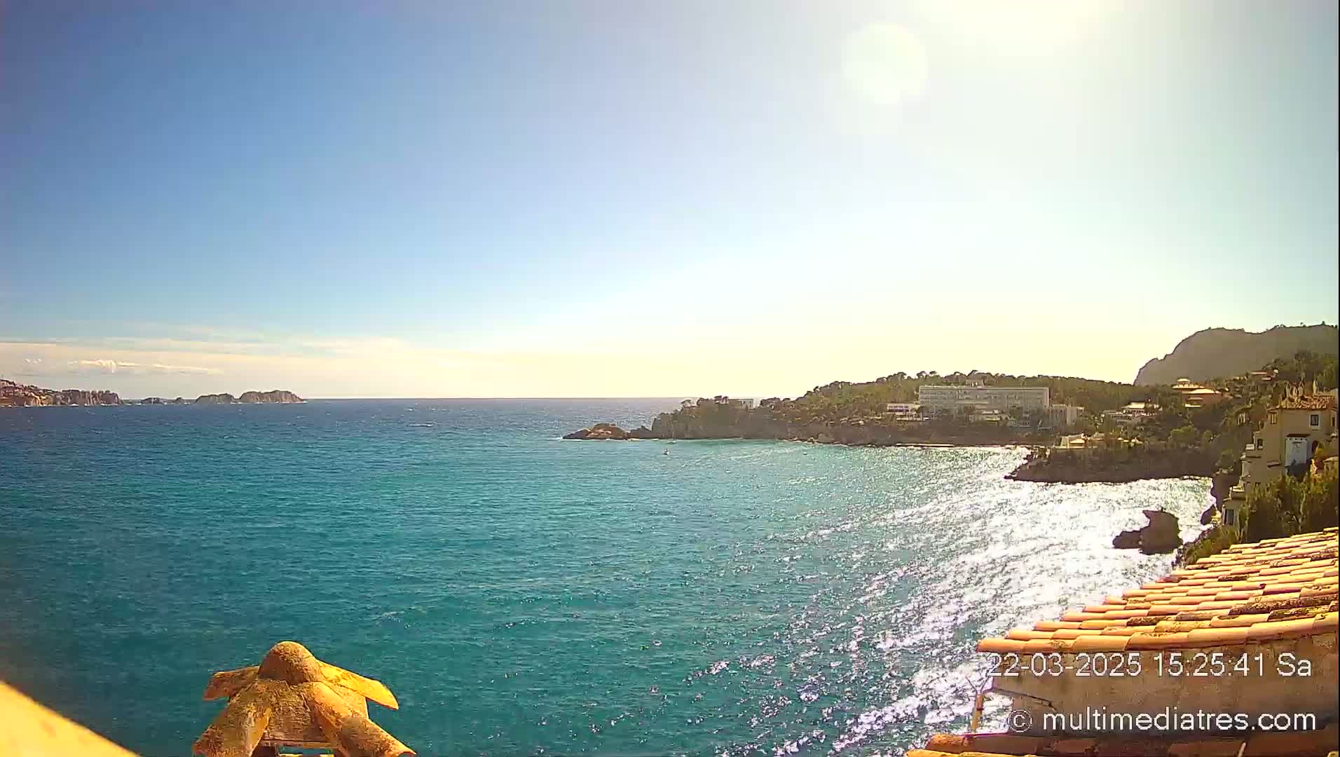 A sunny day reveals a tranquil turquoise sea lapping against a rocky coastline, dotted with buildings and distant islands under a clear sky.