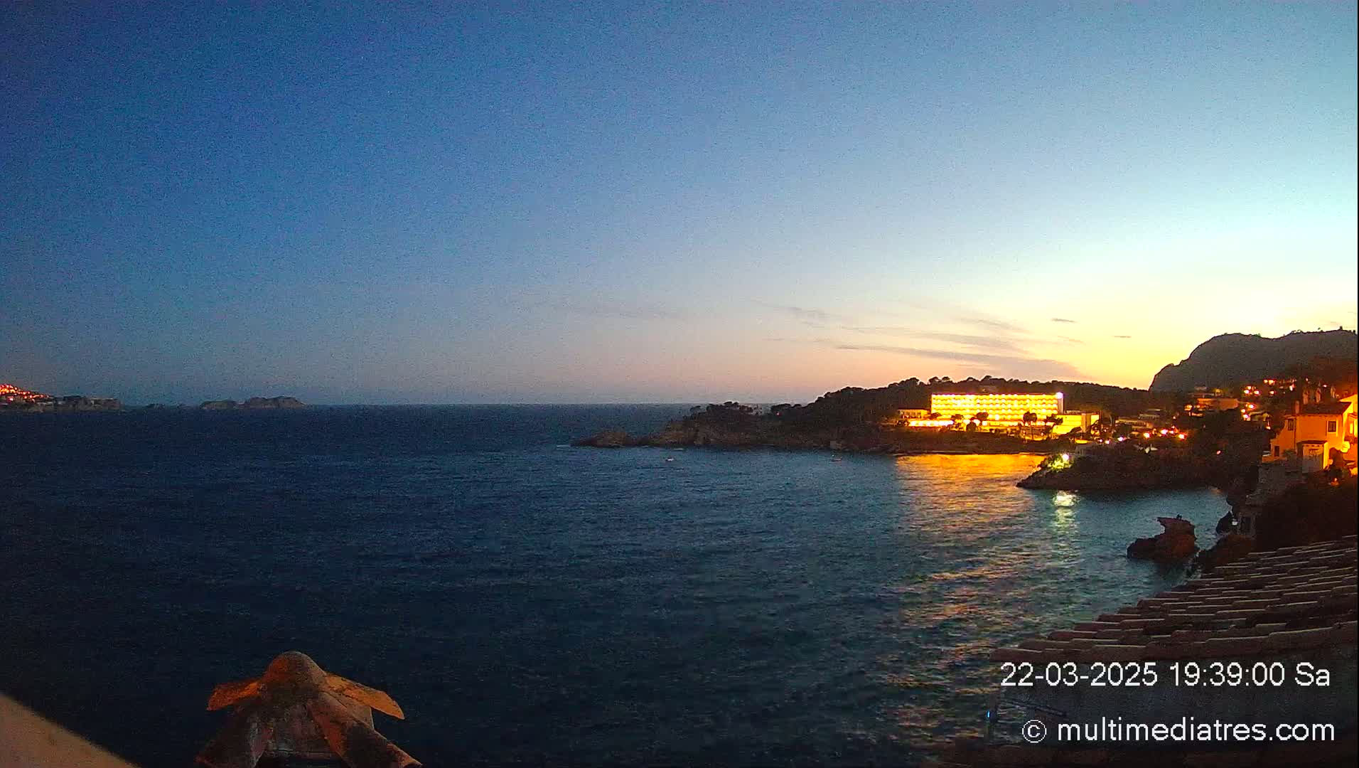 A coastal scene at dusk shows a calm, dark-blue sea with a brightly lit hotel and buildings on a shoreline, and a hill in the background.