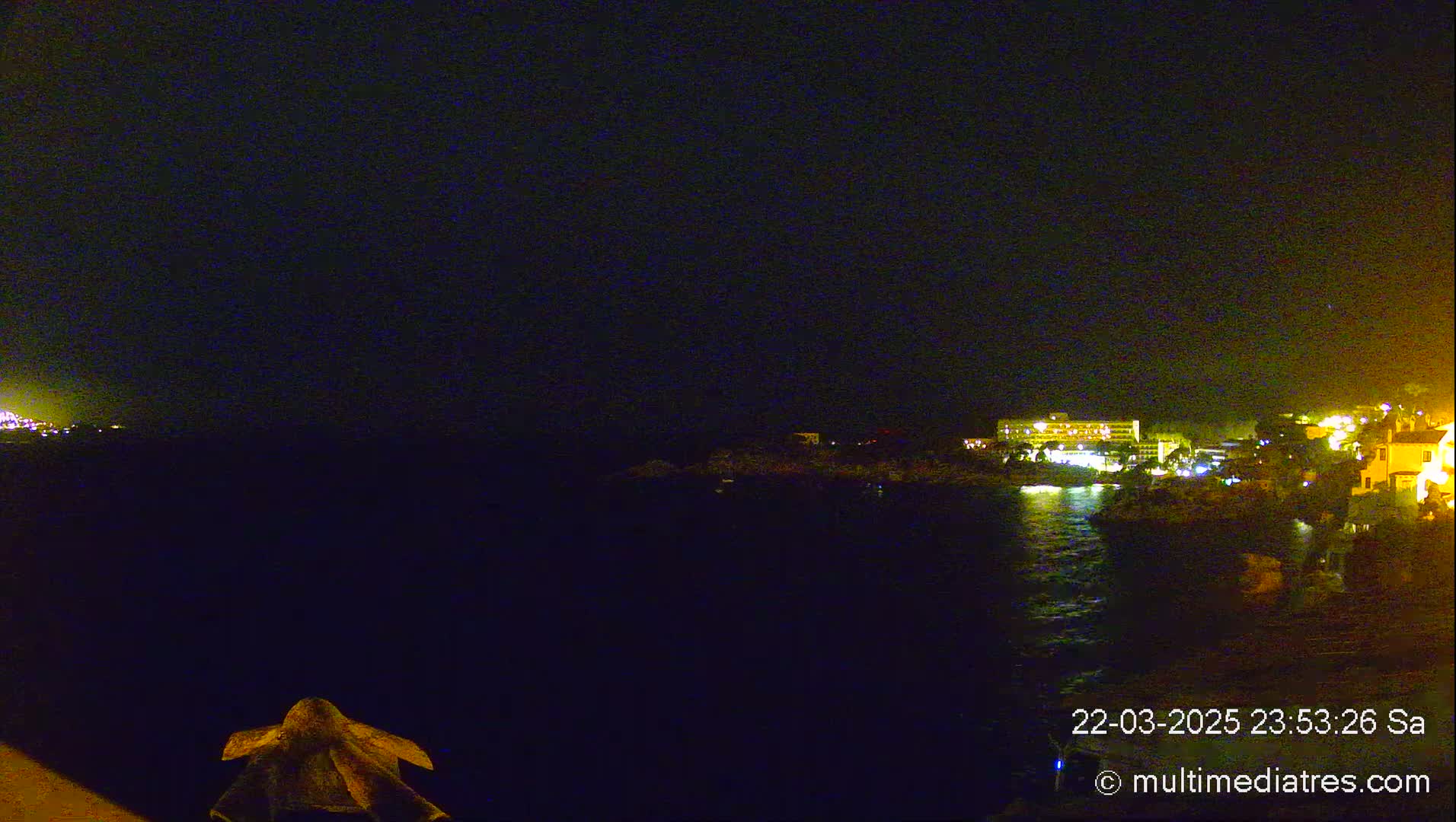A nighttime view of a dark body of water with a distant illuminated shoreline featuring buildings under a clear, dark sky.