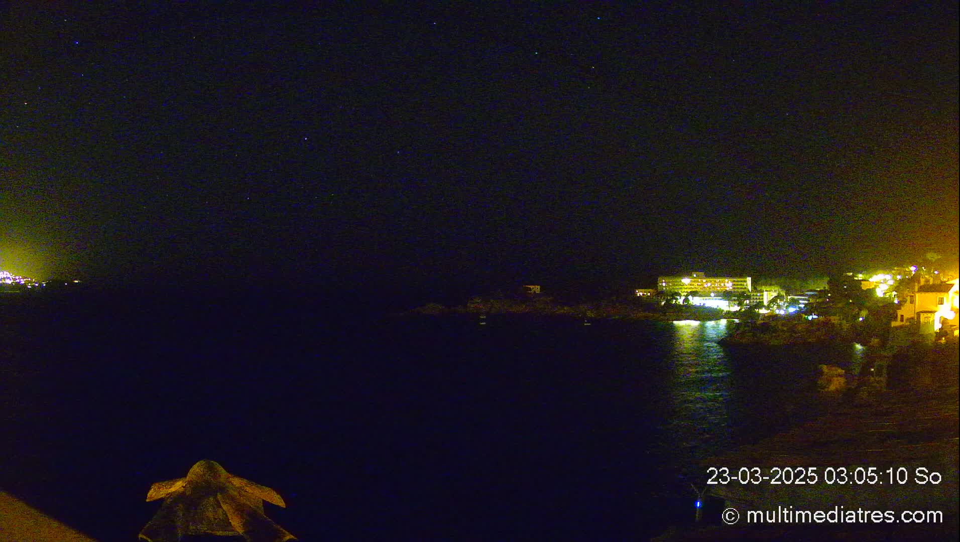 A nighttime coastal scene shows a dark body of water reflecting the lights of buildings on a distant shore under a clear, starlit sky.