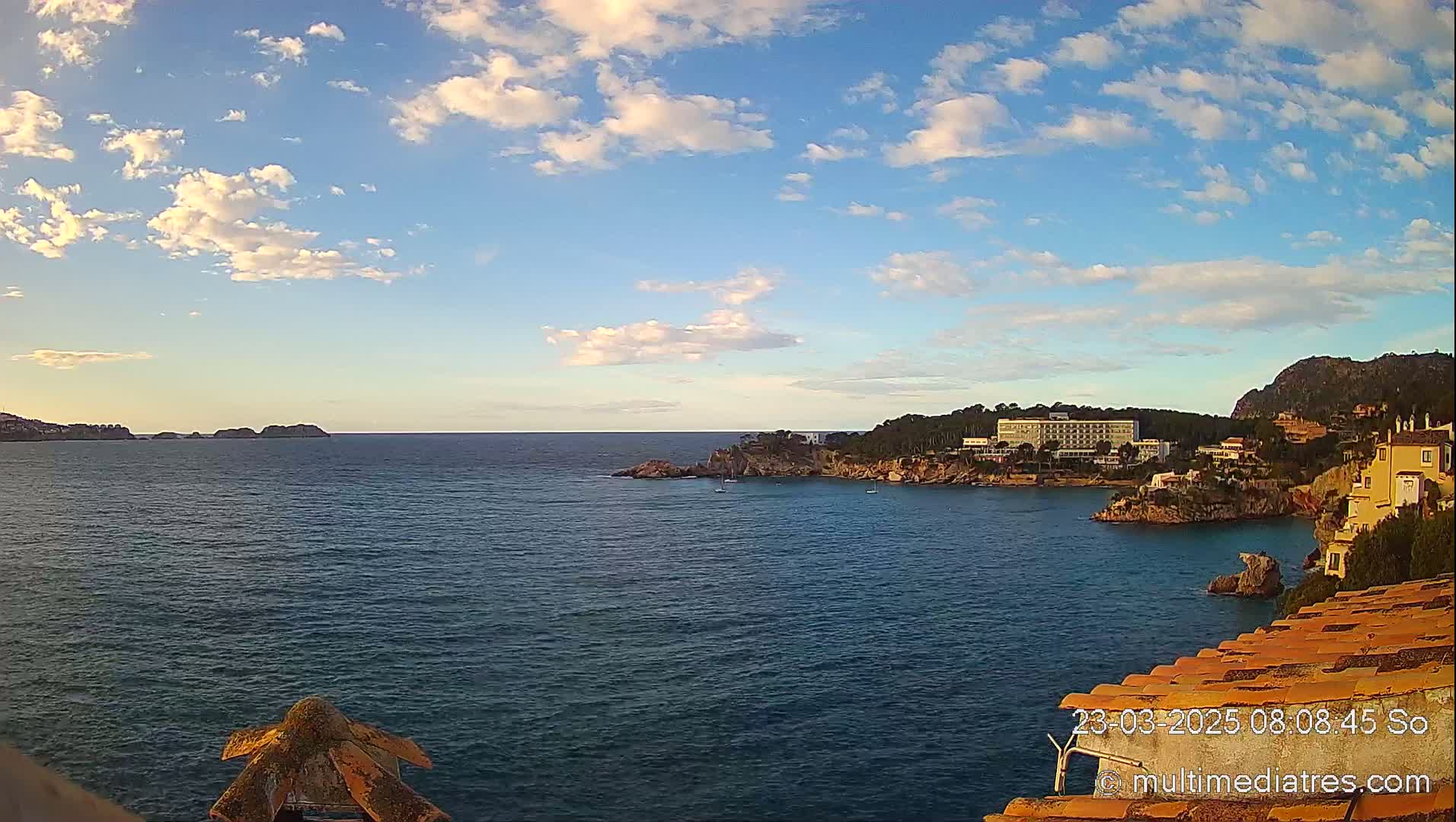 A calm, partly cloudy day shows a coastal scene with a large hotel and other buildings on a rocky hillside overlooking a tranquil bay.