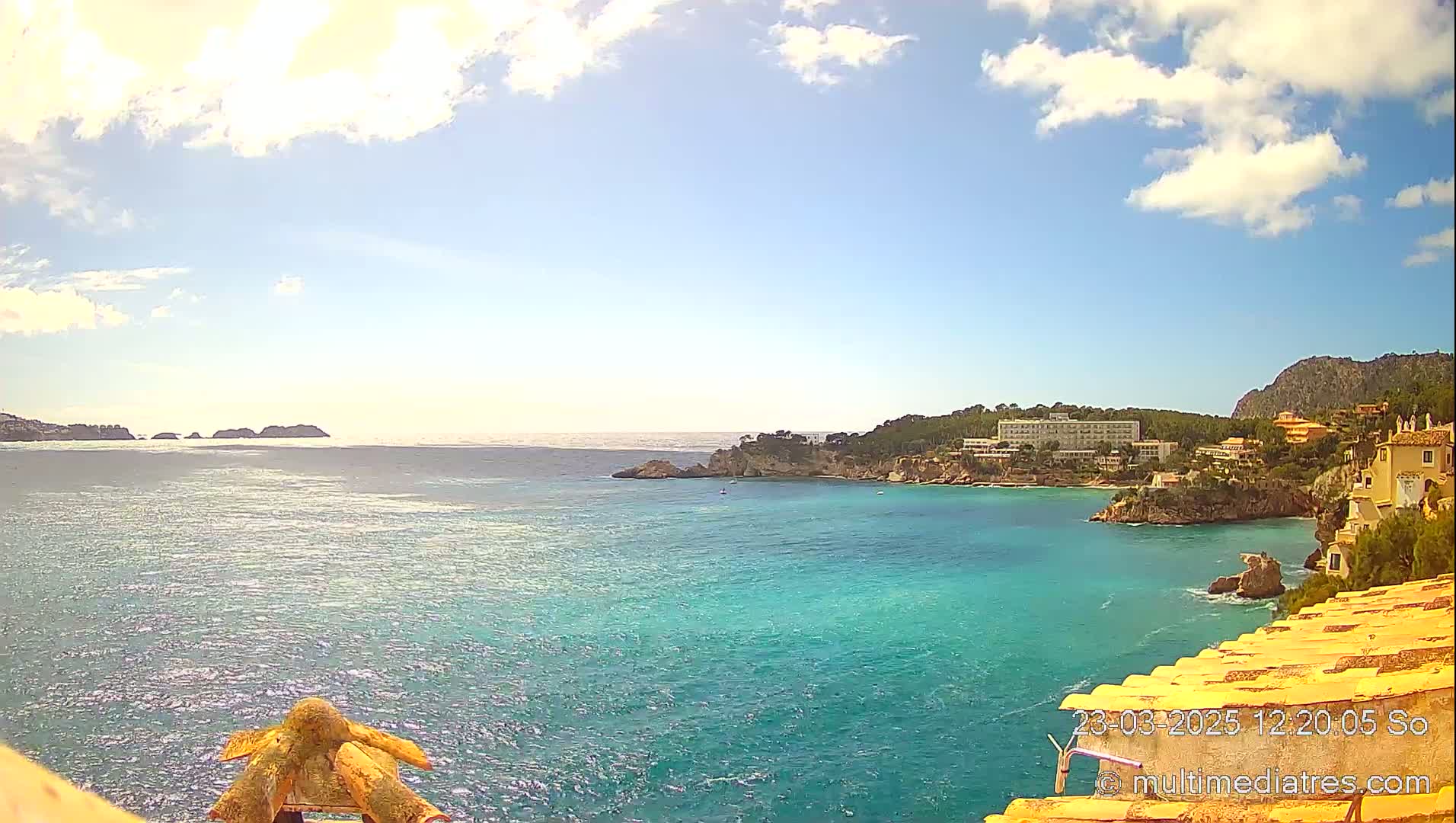 A partly sunny day reveals a tranquil turquoise bay, nestled between rocky headlands, with a hotel and buildings visible along the coast.