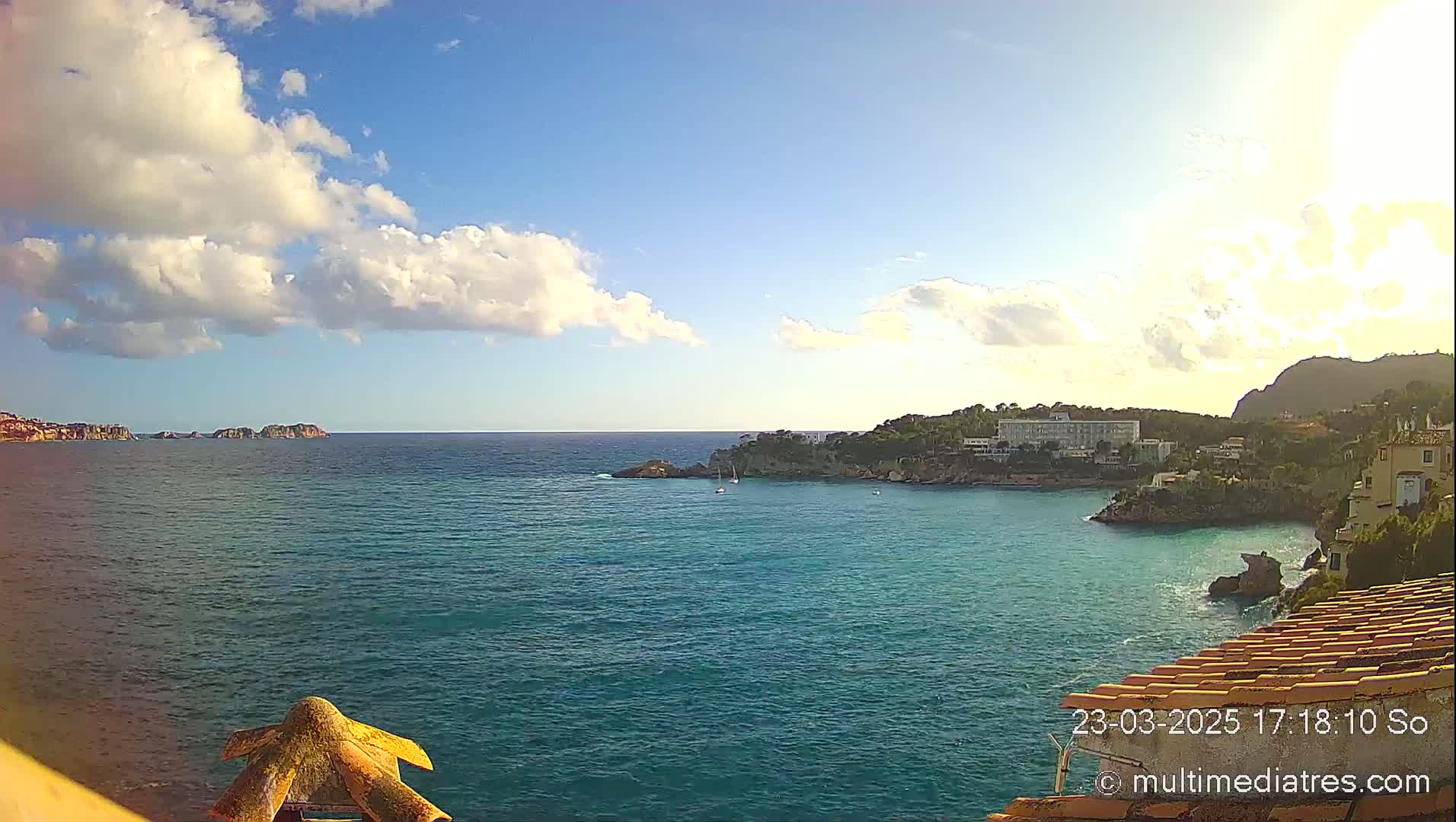 A partly cloudy, sunny day overlooks a calm turquoise bay with a coastline of buildings and vegetation.