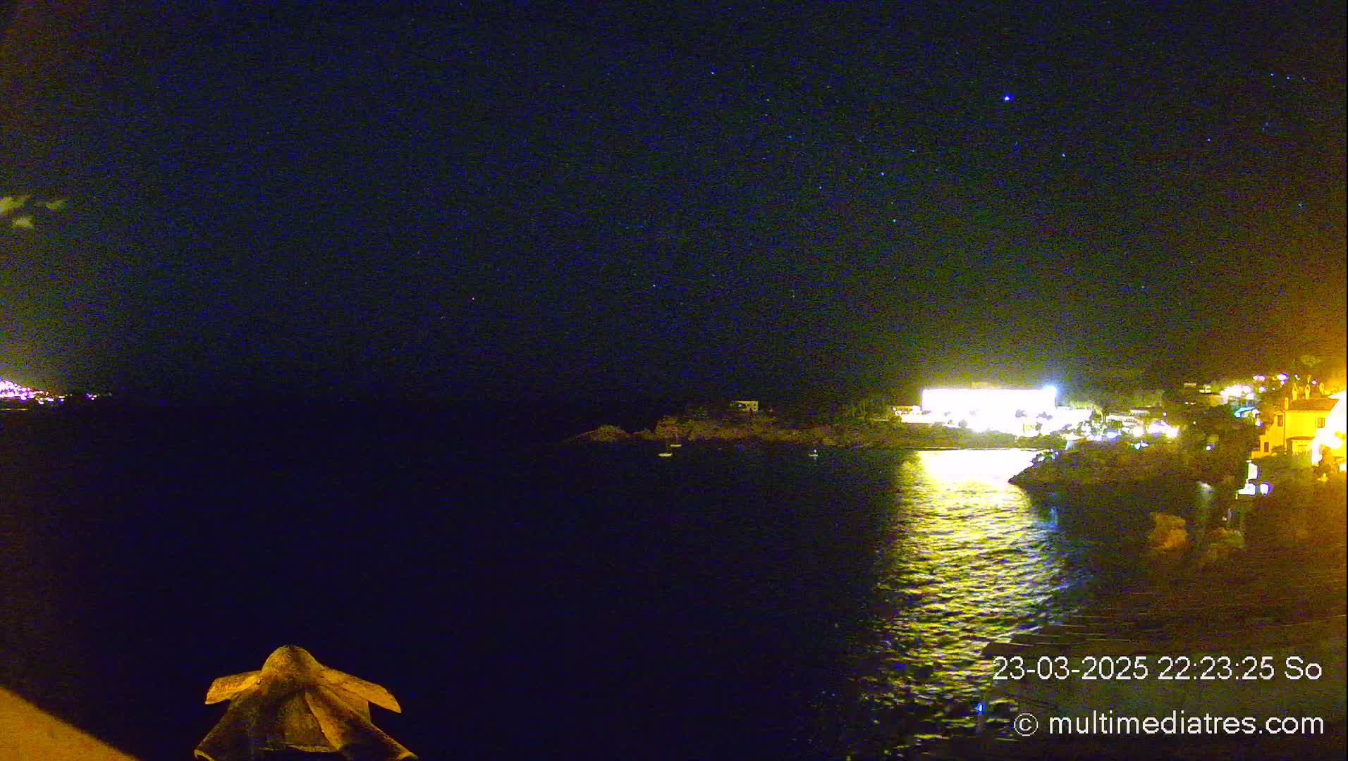 A nighttime view of a calm body of water reflecting the lights of a nearby town under a clear, starlit sky.