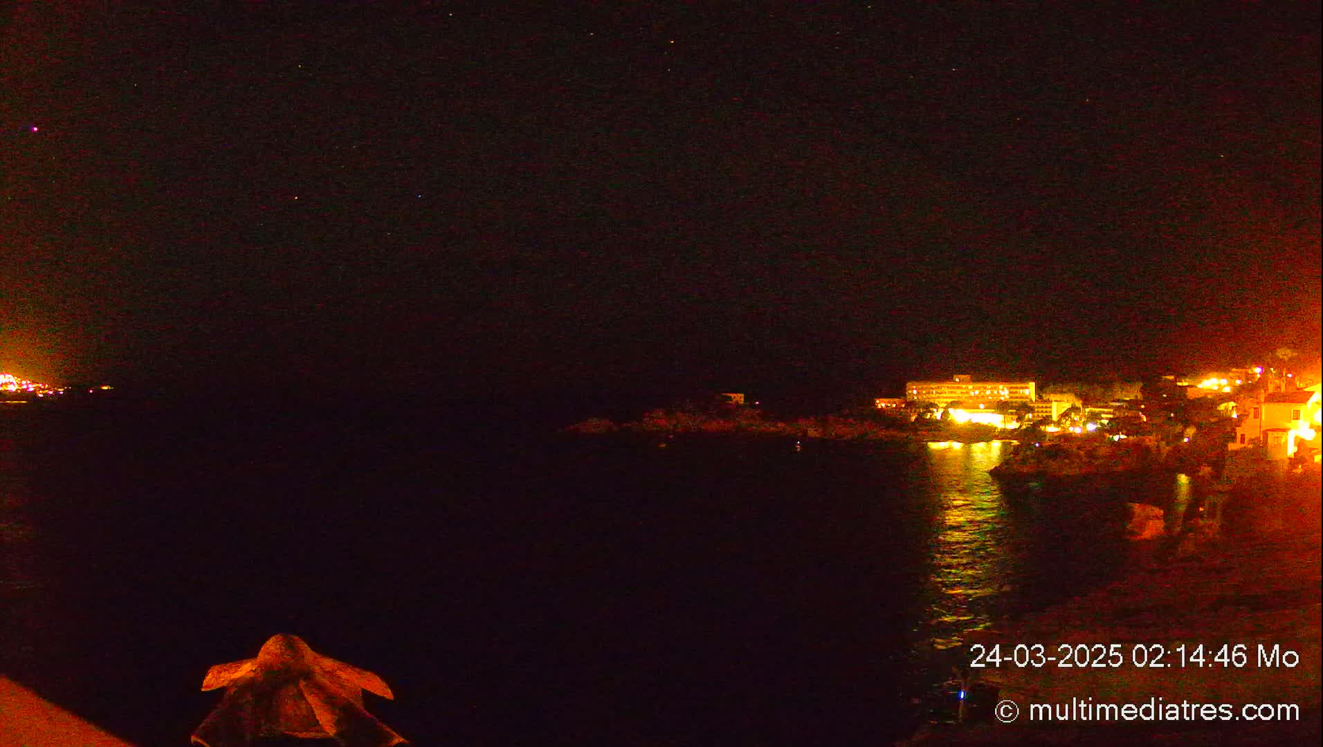 A nighttime view of a dark body of water reflecting the lights of buildings on the shore under a clear, starry sky.