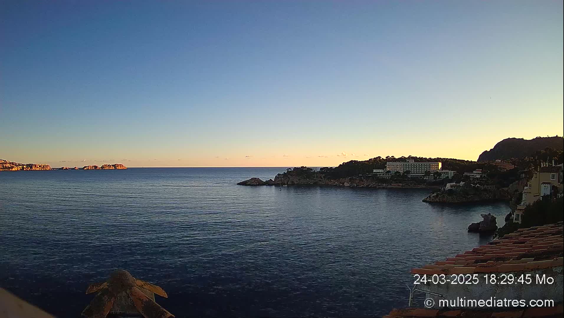 A calm sea at sunset, with a coastline featuring buildings and rocky outcrops under a clear blue sky.