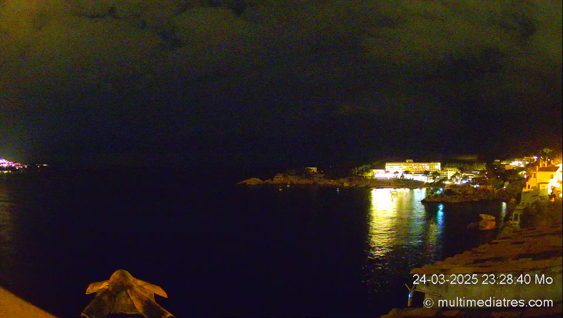 A nighttime view of a calm body of water reflecting the lights of buildings on a distant shore under a dark, cloudy sky.