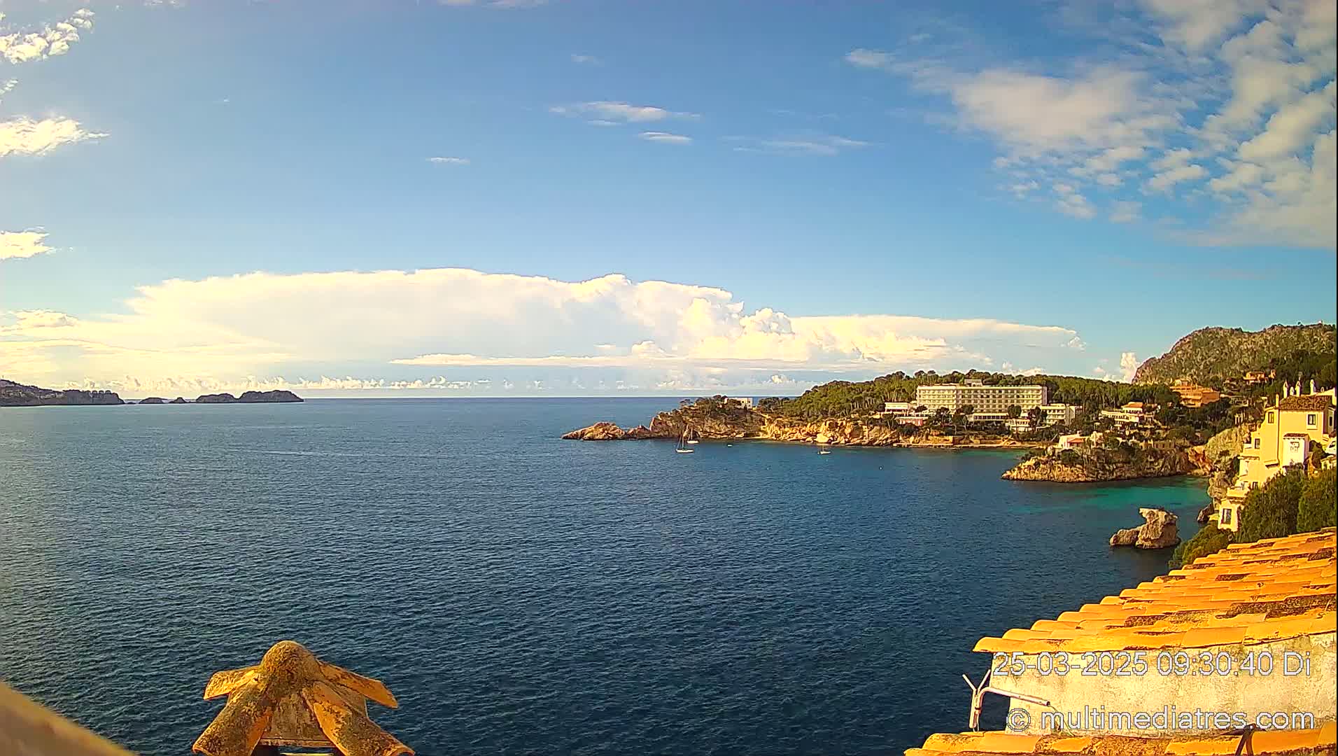 A partly cloudy sky overlooks a calm, blue sea with a coastline featuring buildings, trees, and rocky outcrops.