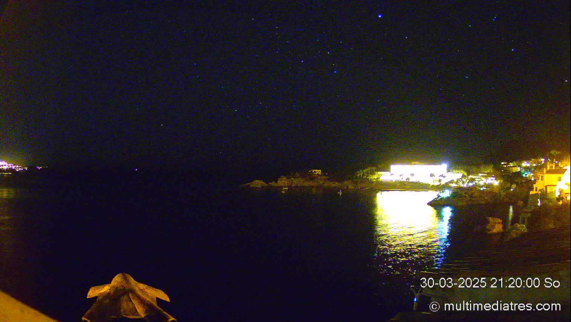 A nighttime view of a calm body of water reflecting the lights of buildings on the shore under a clear, starry sky.