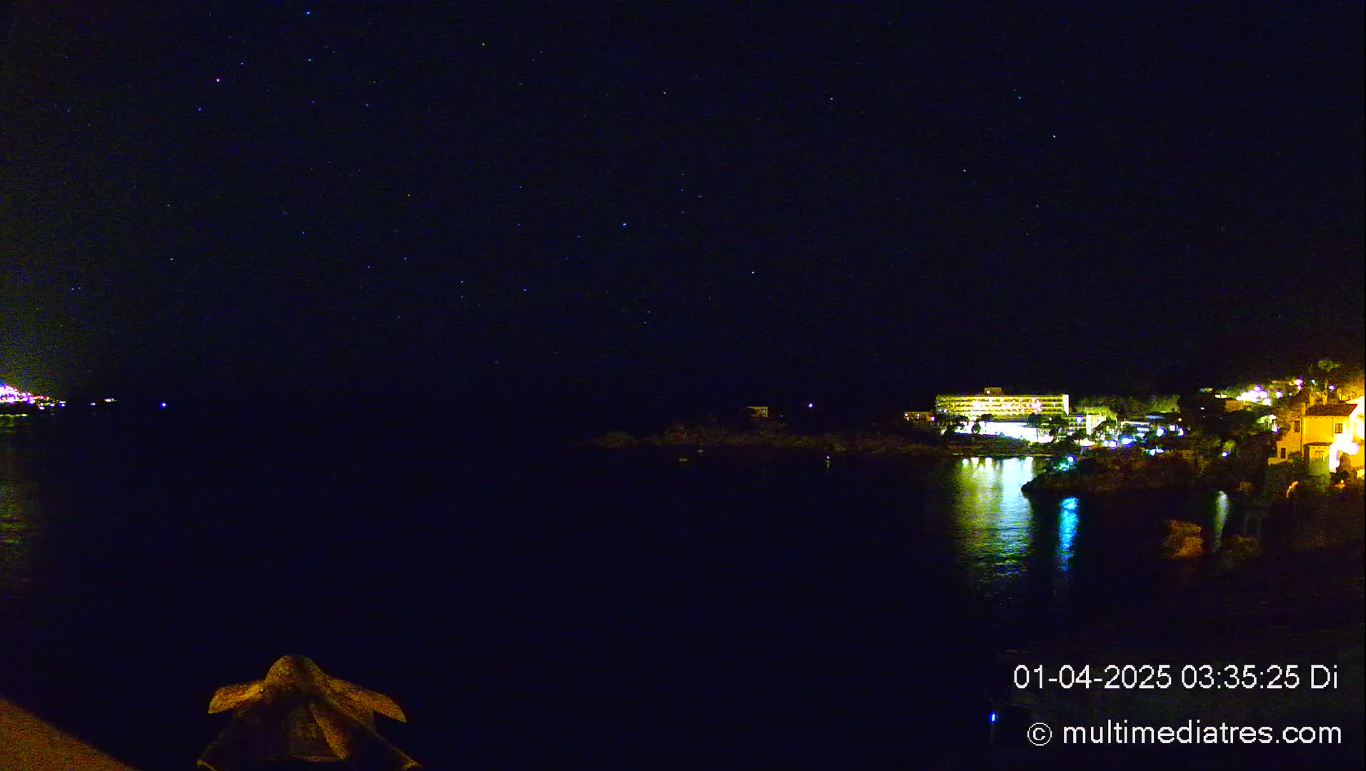 A dark, starlit night scene shows a calm body of water reflecting the lights of buildings on the opposite shore.