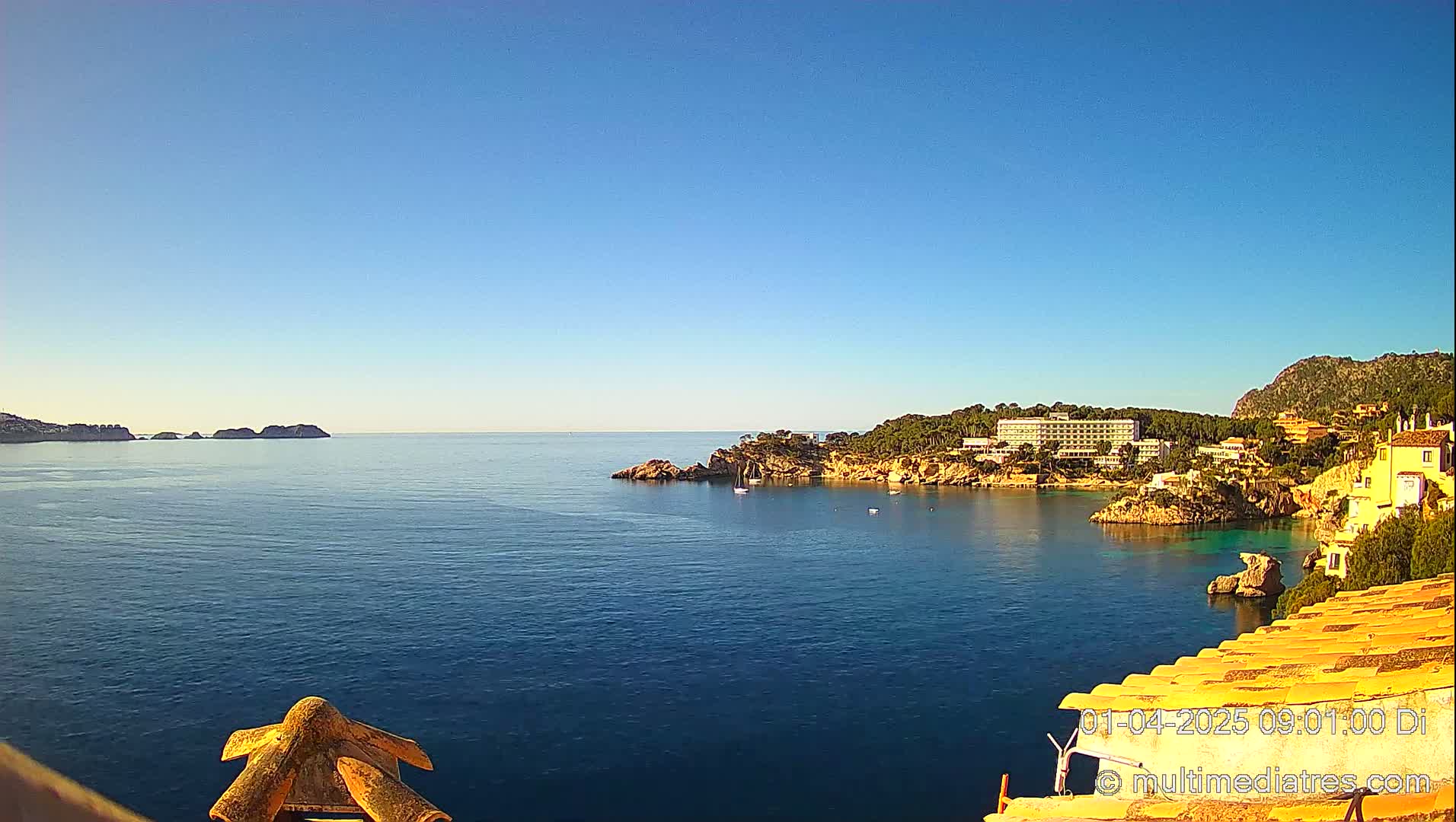 A tranquil coastal scene on a sunny day shows a calm blue sea, a rocky shoreline with buildings and trees, and distant islands.