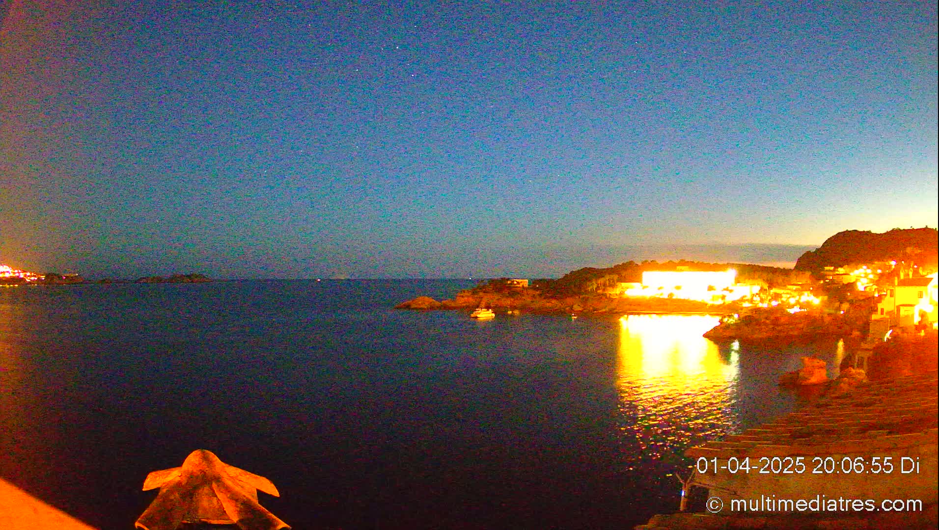 A nighttime coastal scene shows a dark body of water reflecting the lights of buildings on a nearby shore under a clear, starry sky.