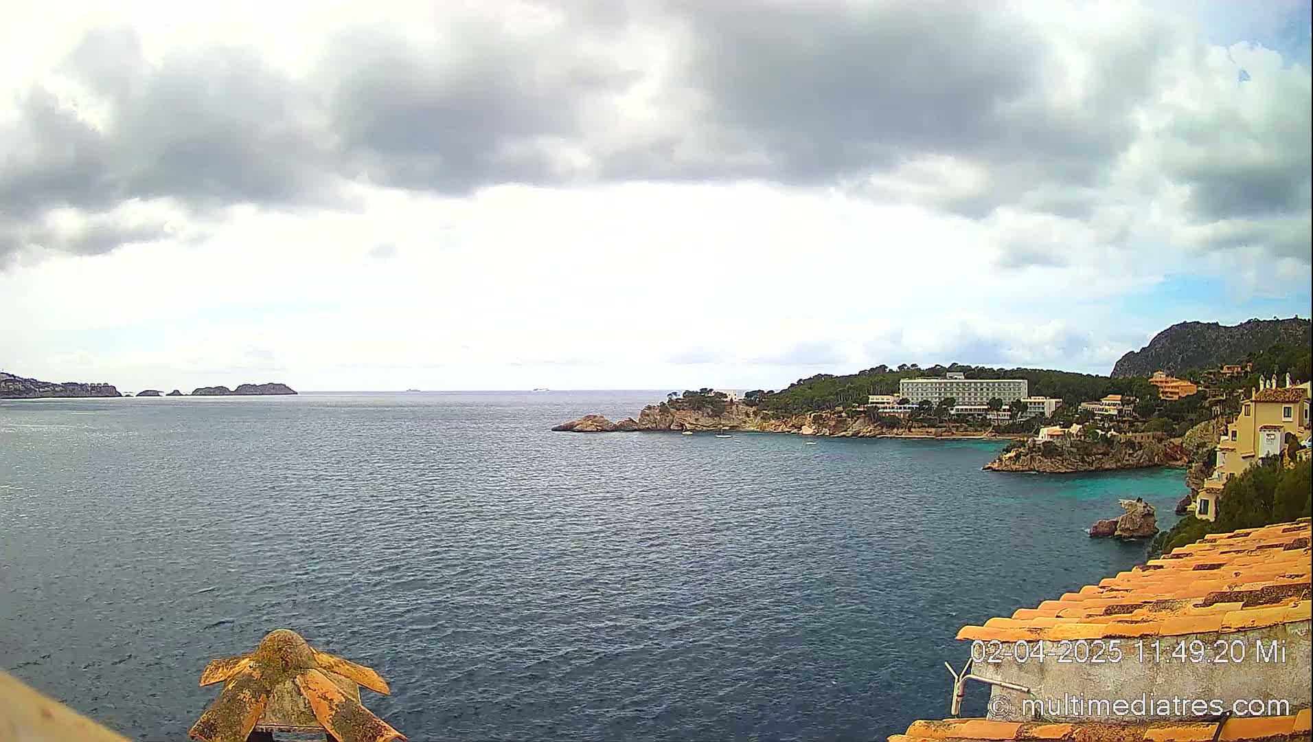 A coastal scene shows a calm sea under a cloudy sky, with buildings and vegetation on a rocky shoreline and small islands in the distance.