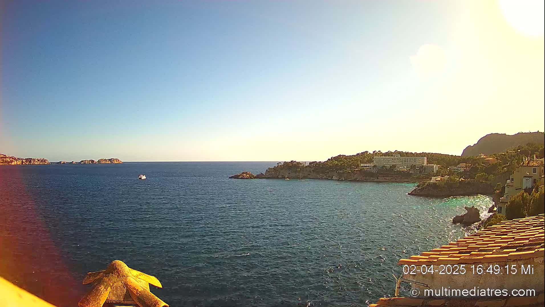 A clear, sunny day overlooking a calm sea with a coastline featuring buildings and vegetation, and a small boat in the distance.