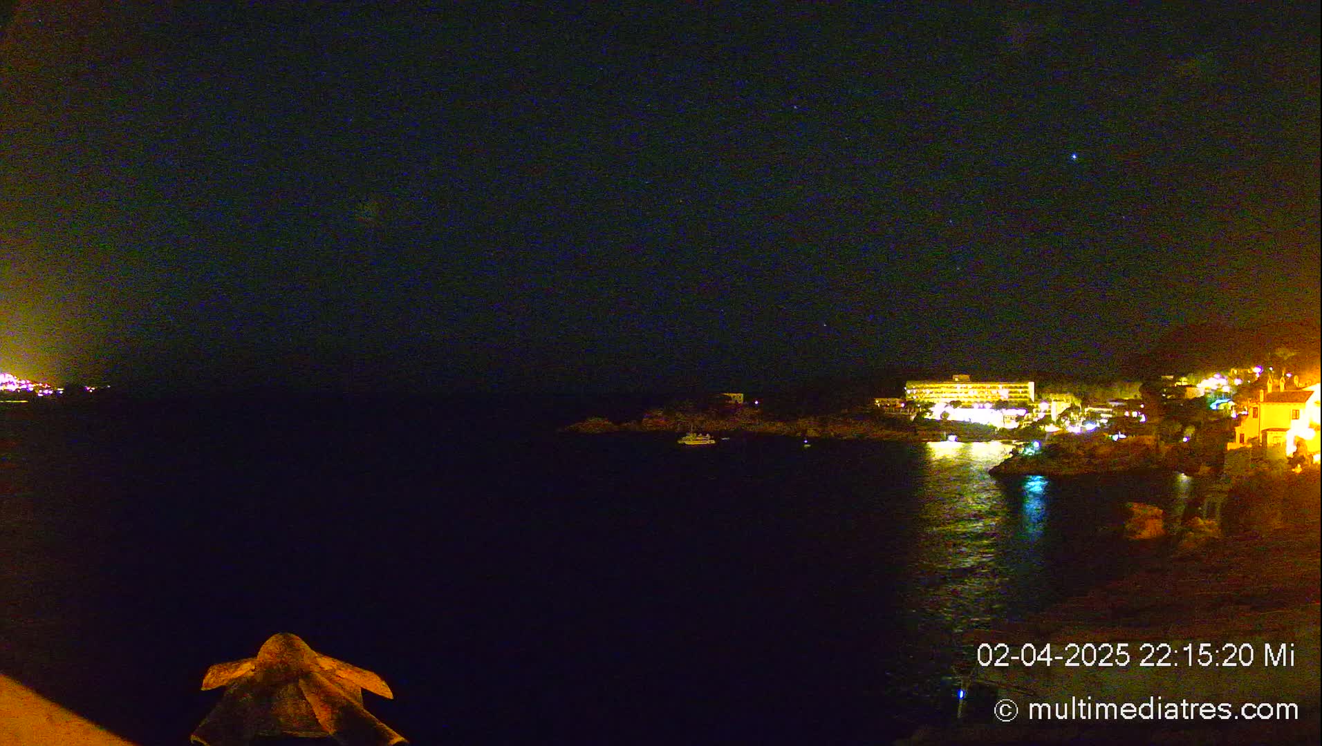 A nighttime view of a calm body of water with illuminated buildings and a small boat on the shore under a clear, starry sky.