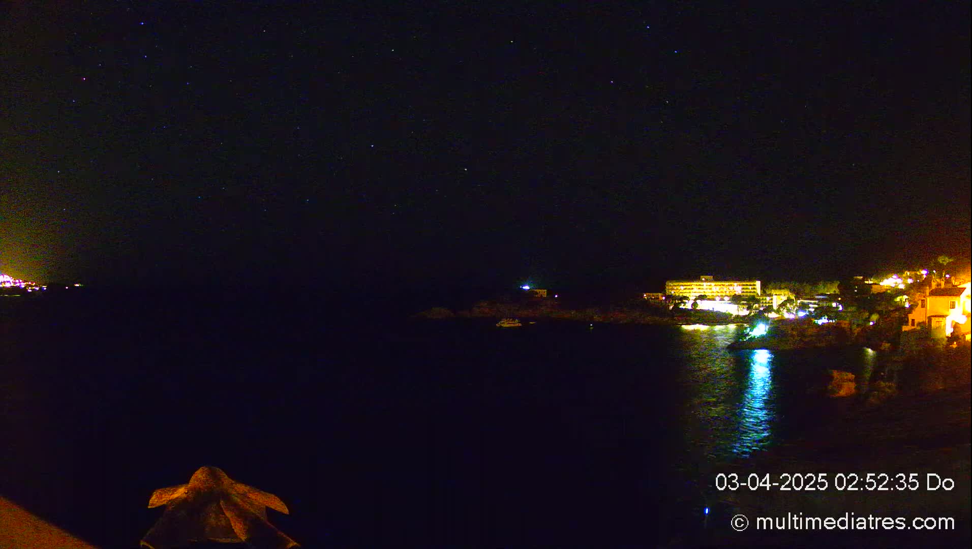 A nighttime seaside view shows a starlit sky over dark water reflecting the lights of buildings on a distant shore.