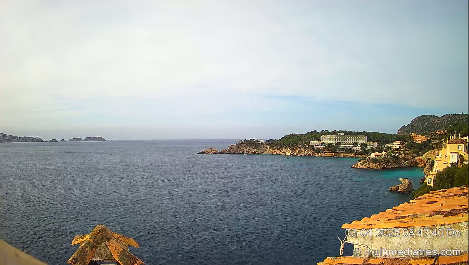 A coastal scene shows a calm ocean under a mostly cloudy sky, with a hotel and other buildings nestled on a rocky shoreline.