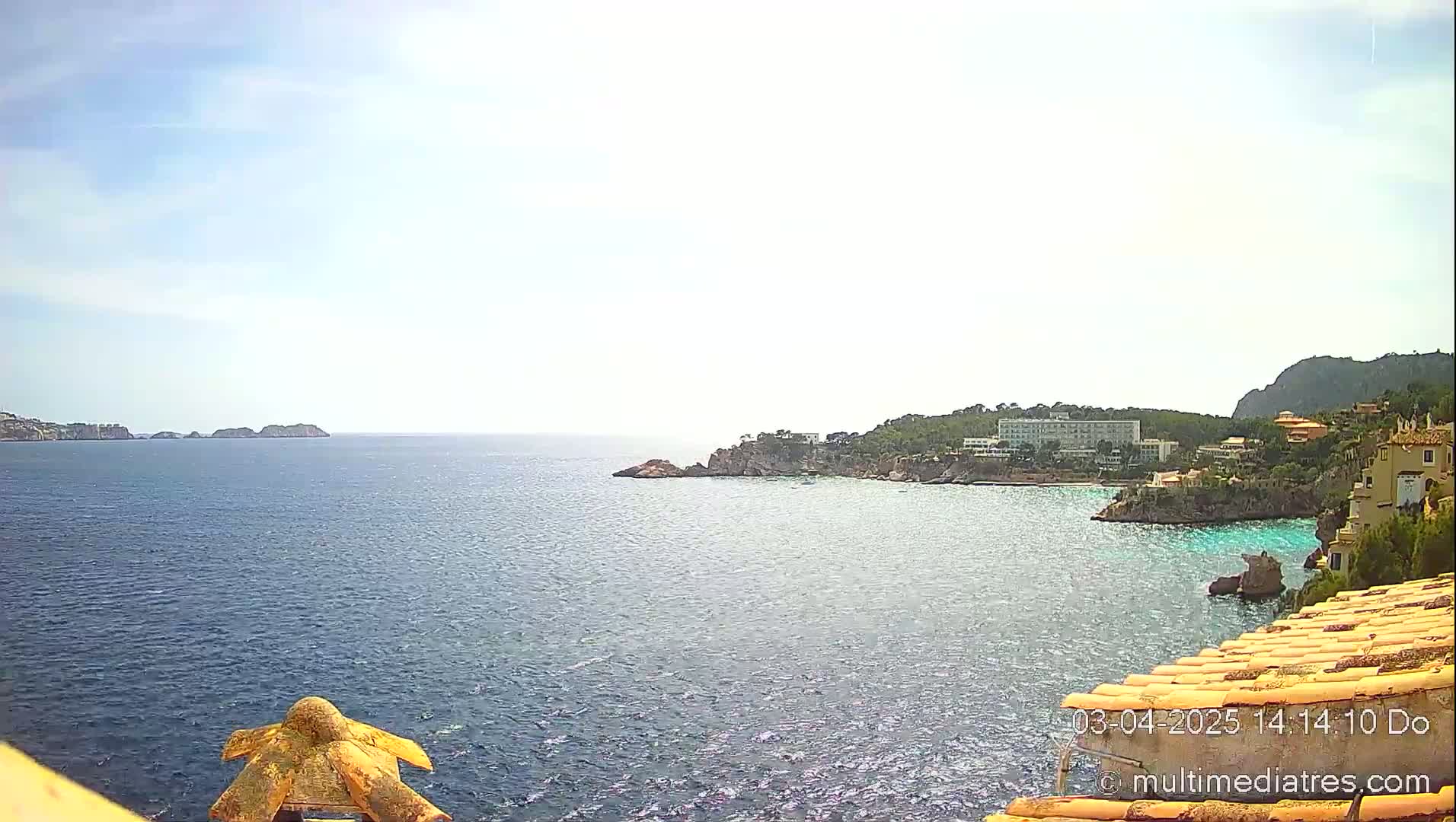 A coastal scene on a partly sunny day shows a calm blue sea with a rocky shoreline featuring buildings and vegetation.