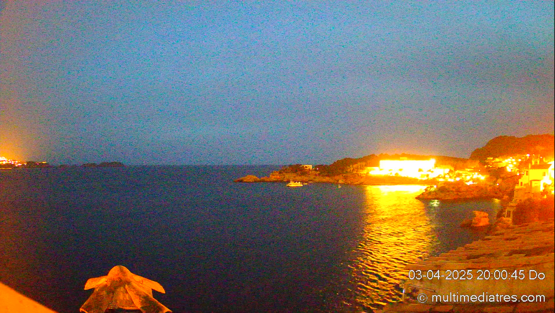 A nighttime view of a calm body of water reflecting the warm, golden lights of buildings on a hill, under a twilight sky.