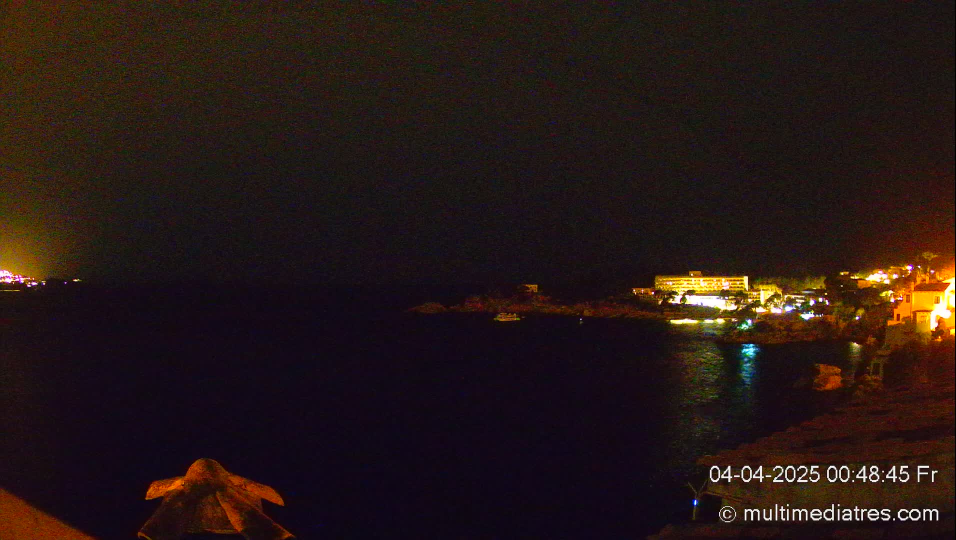 A nighttime view of a calm body of water with illuminated buildings and vegetation on the opposite shore under a dark sky.