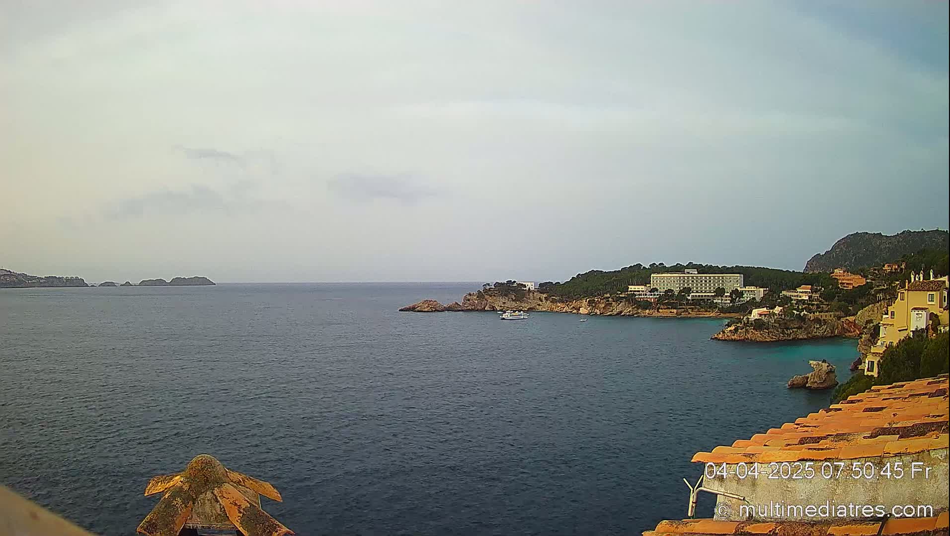 A calm, overcast day reveals a coastal scene with a hotel and buildings nestled into a rocky cove, overlooking a dark-blue sea and distant islands.