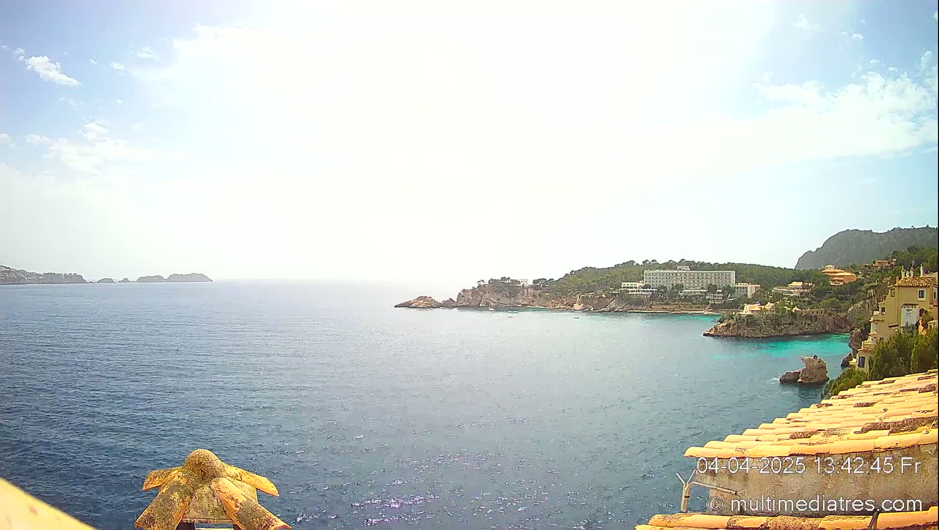 A sunny day shows a coastal view of a calm blue sea with rocky shorelines, buildings, and trees, including a large building on a prominent headland.