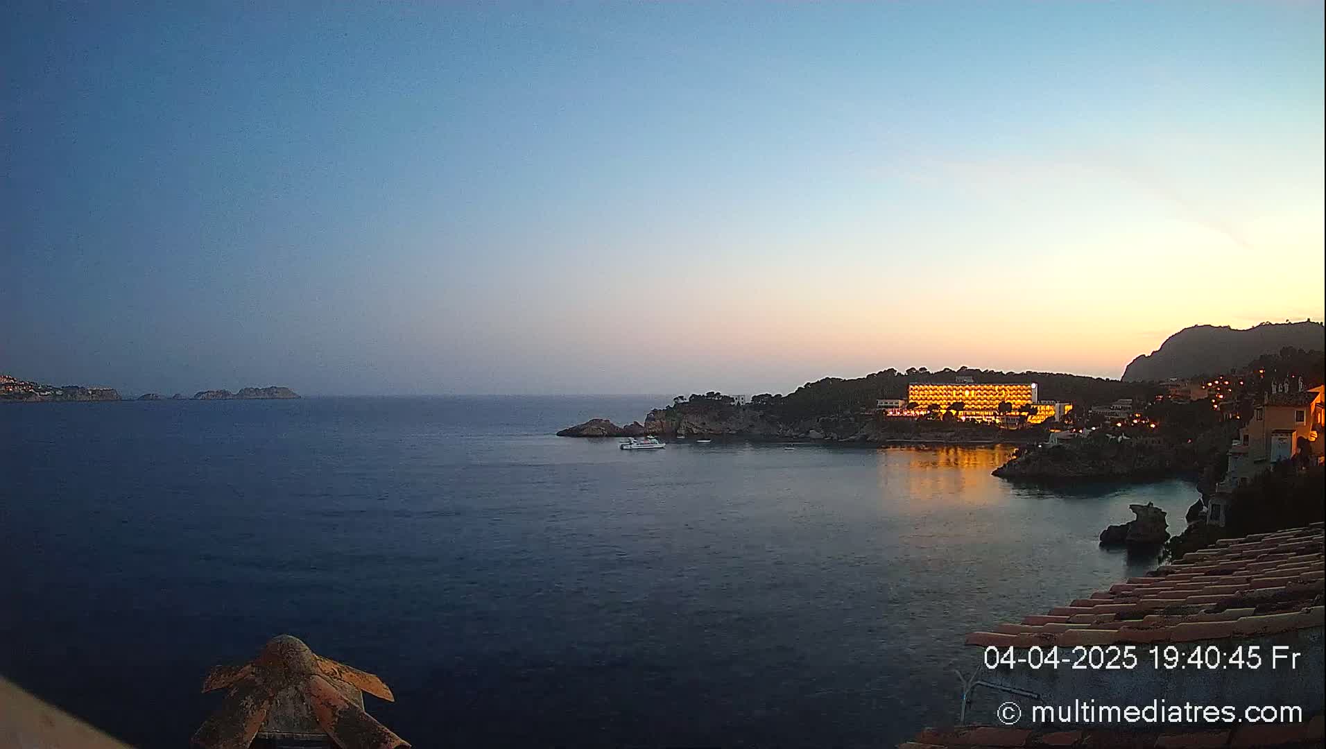 A twilight coastal scene shows a calm sea, a brightly lit hotel on a rocky shoreline, and small islands in the distance under a clear sky.
