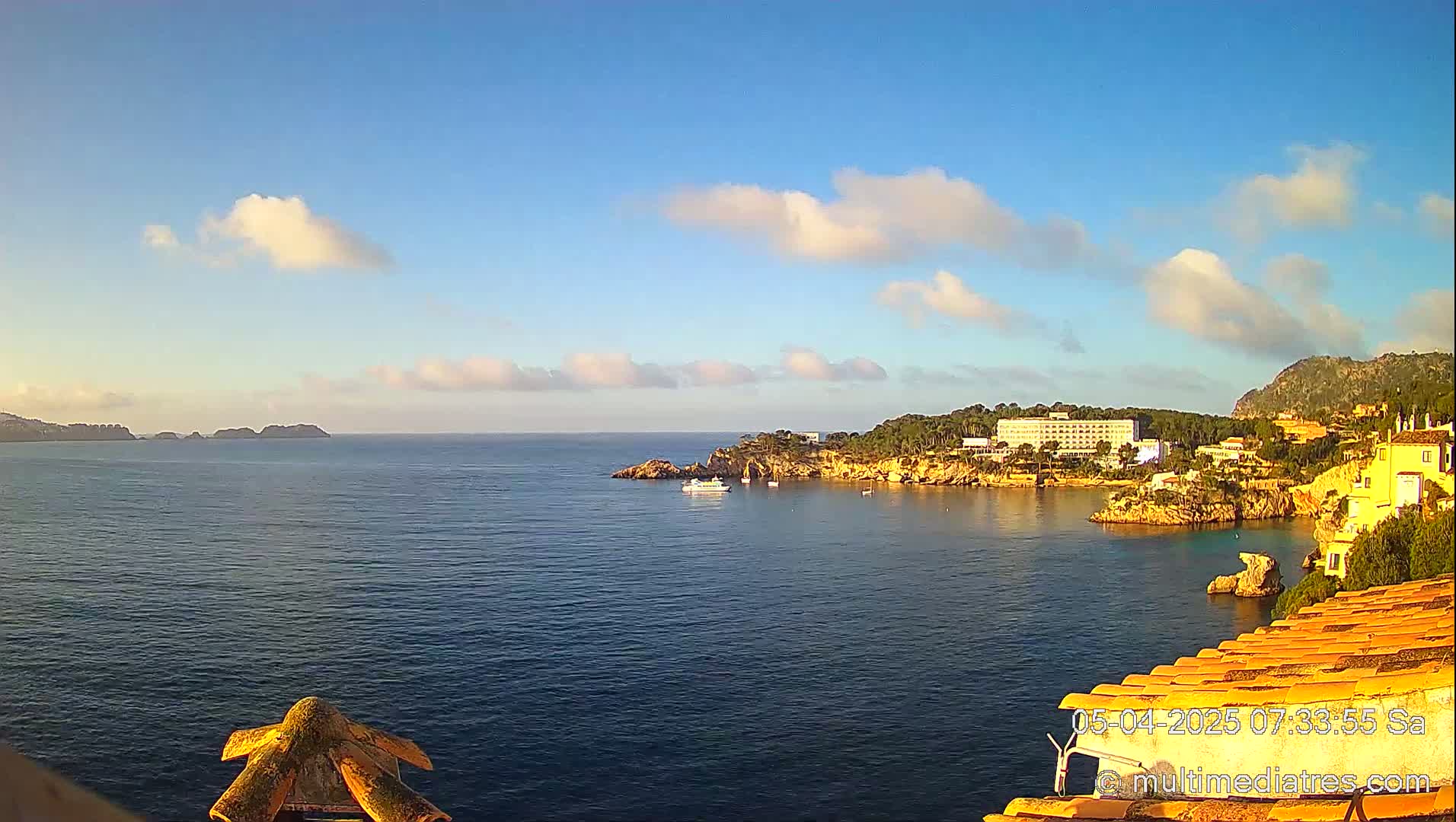 A calm, blue ocean bay is seen from an elevated vantage point, under a mostly sunny sky with a few scattered clouds, featuring a coastline with buildings and vegetation.