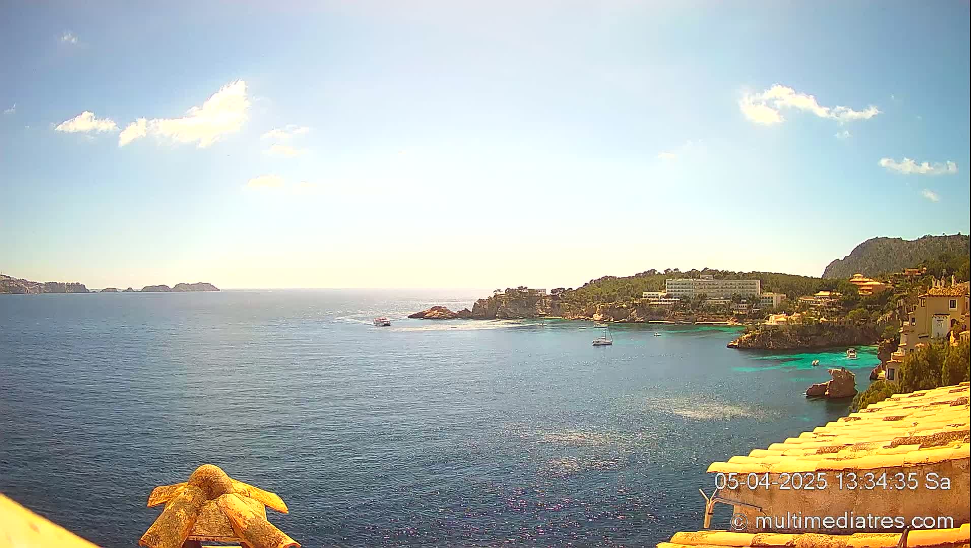 A sunny day reveals a coastal scene with turquoise water, small boats, and buildings nestled on a hillside overlooking a calm bay.