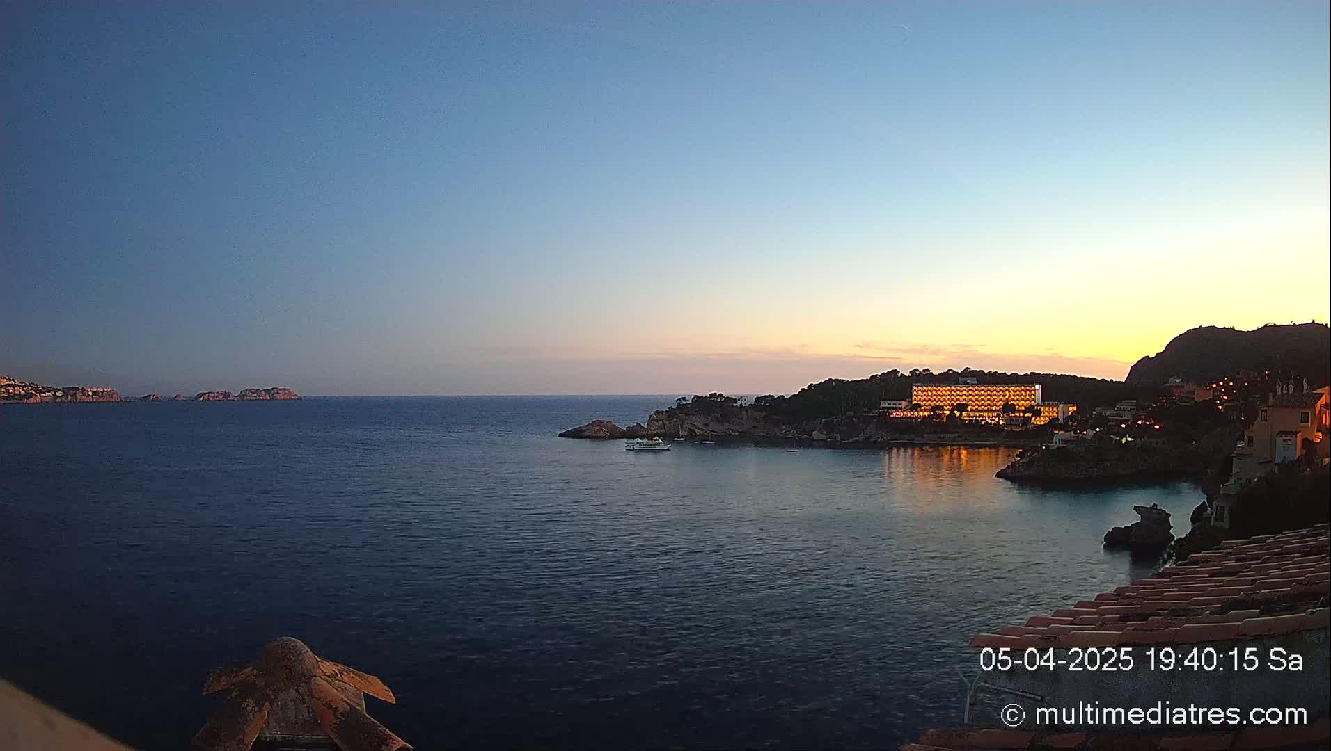A calm, twilight sea scene shows a brightly lit hotel on a rocky shore, with a few small boats in the water and islands in the distance under a clear sky.