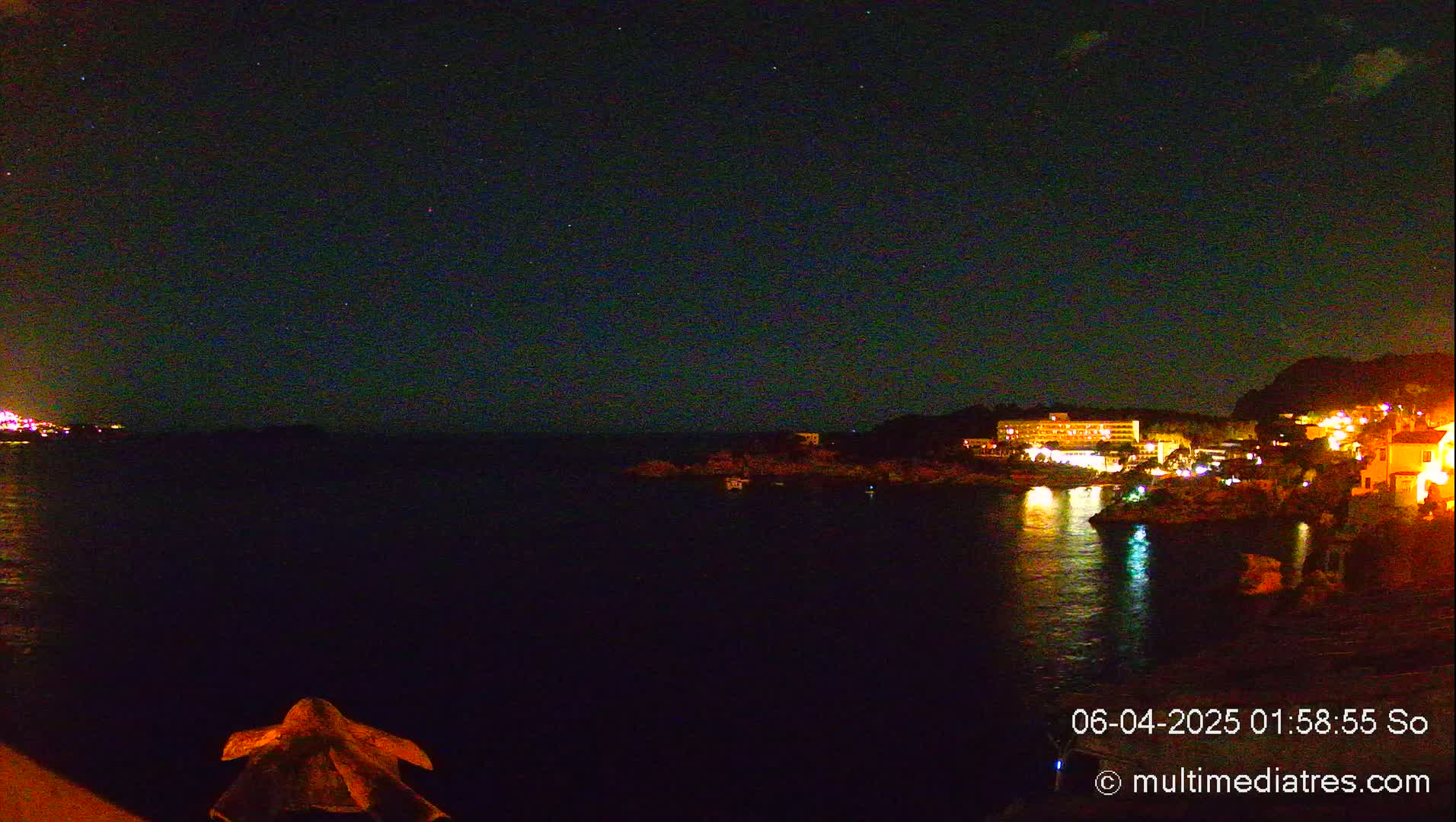 A nighttime view of a calm body of water with a shoreline featuring illuminated buildings under a clear, starry sky.