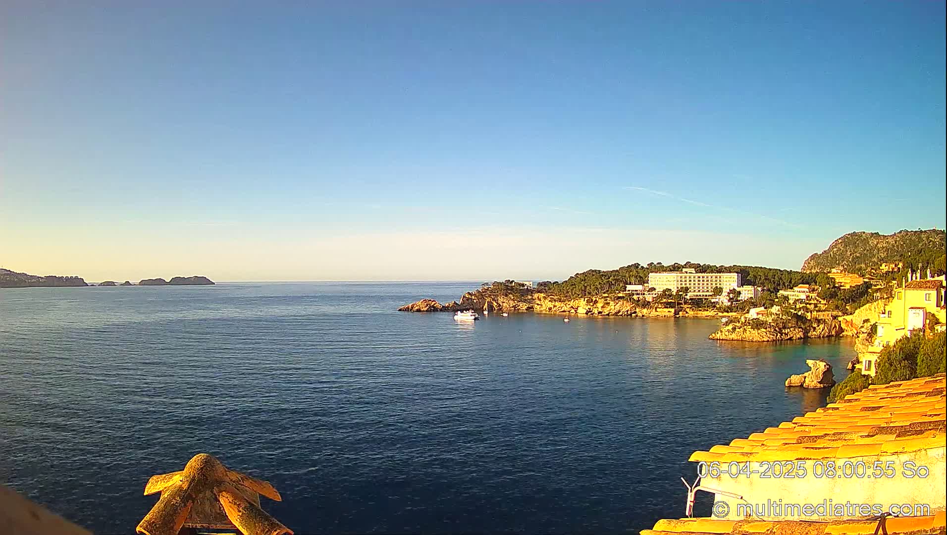 A calm, clear blue sea stretches out from a rocky coastline, featuring a hotel and buildings nestled among trees under a bright, sunny sky.