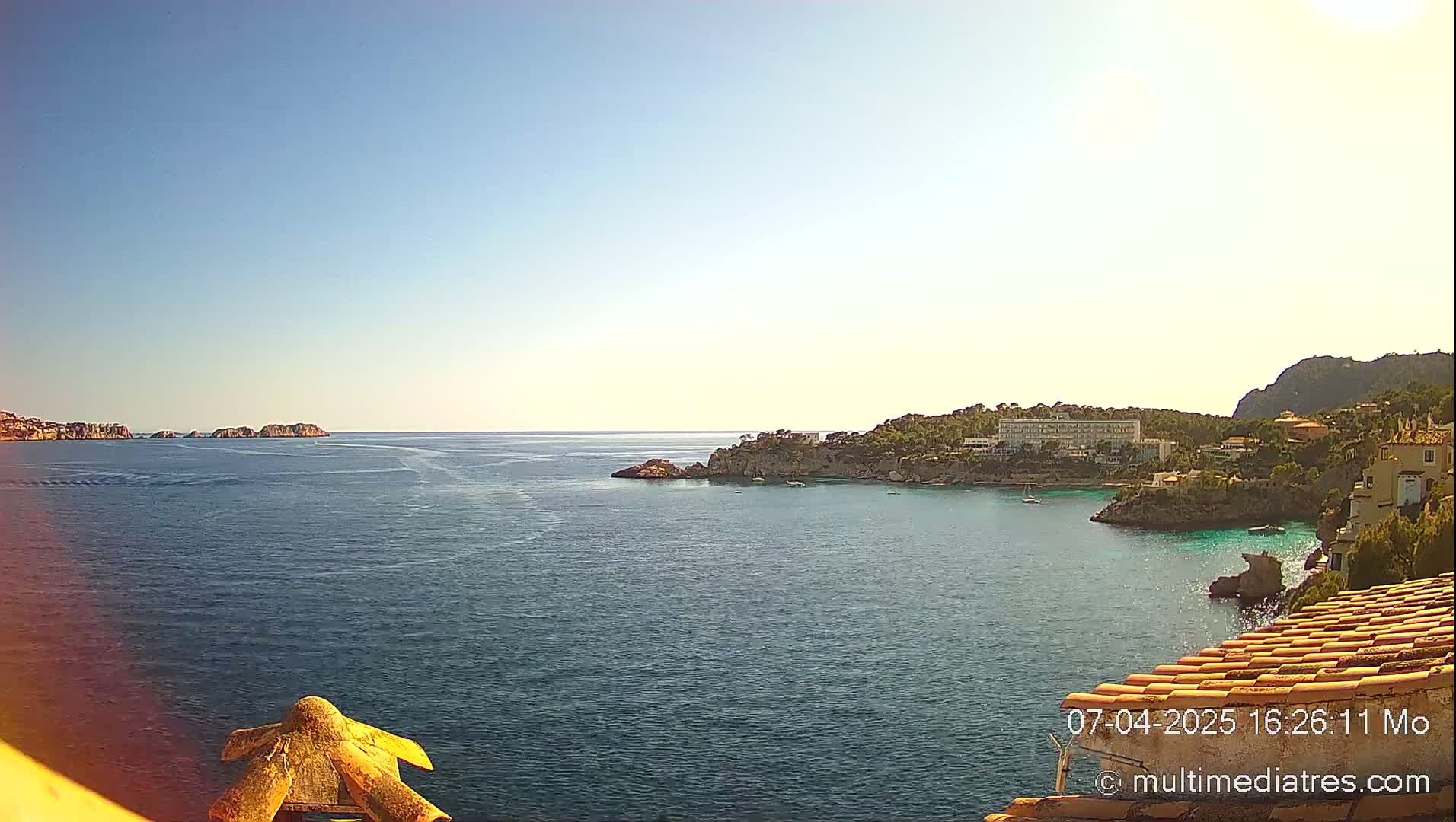 A clear, sunny day overlooks a calm bay with rocky shores, buildings, and some small boats.
