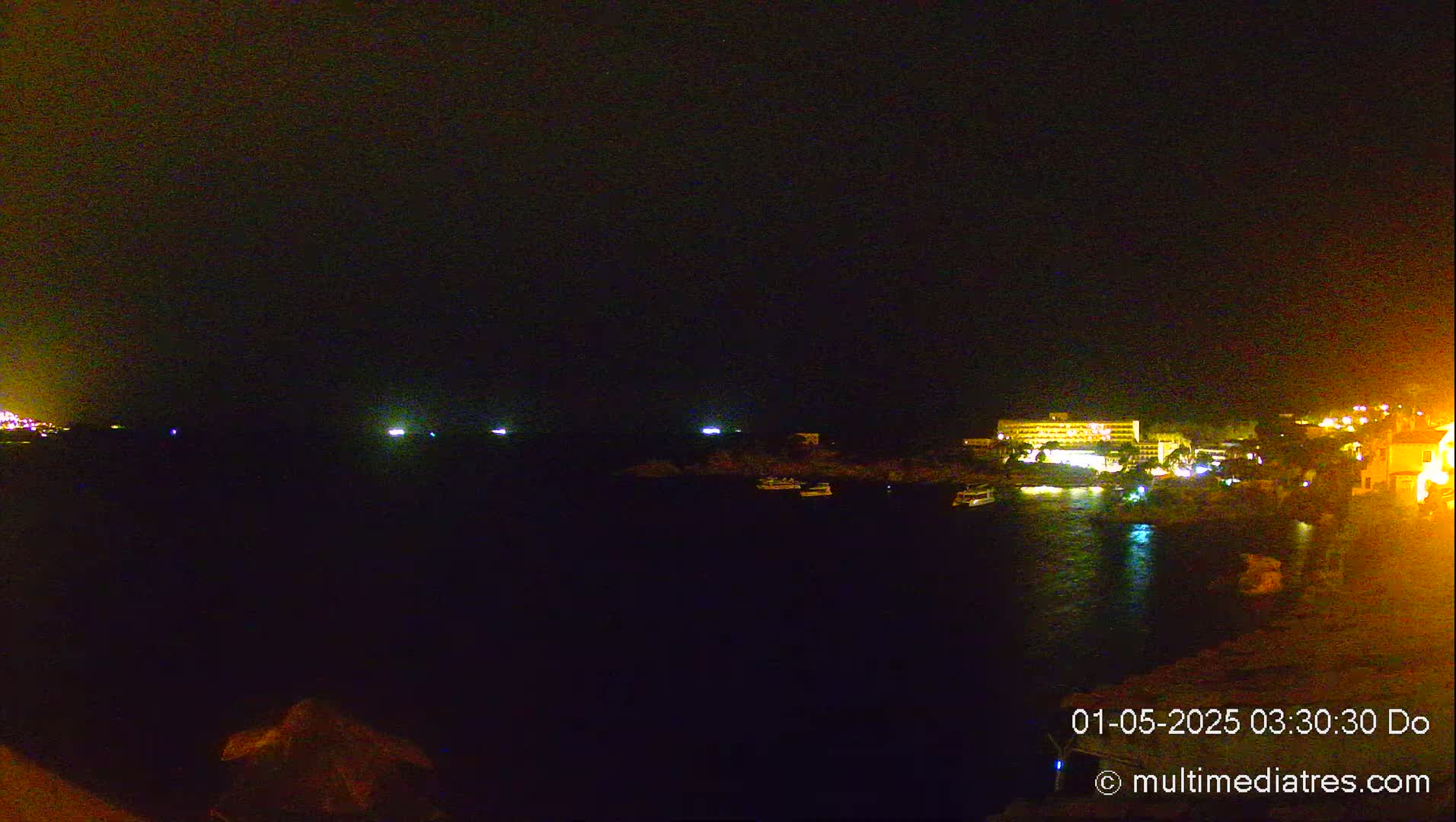 A nighttime view of a dark body of water with faintly lit boats and a brightly lit building on the shore under a dark sky.