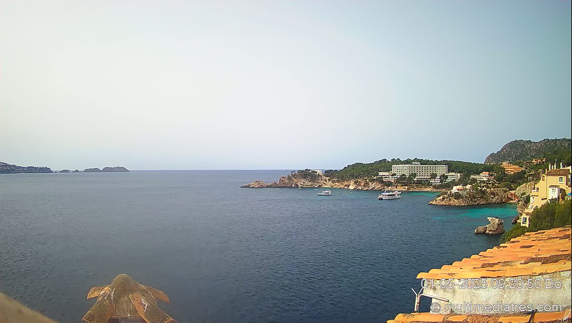 A calm, hazy day reveals a coastal scene of a blue ocean, rocky shores with buildings and trees, and several boats.