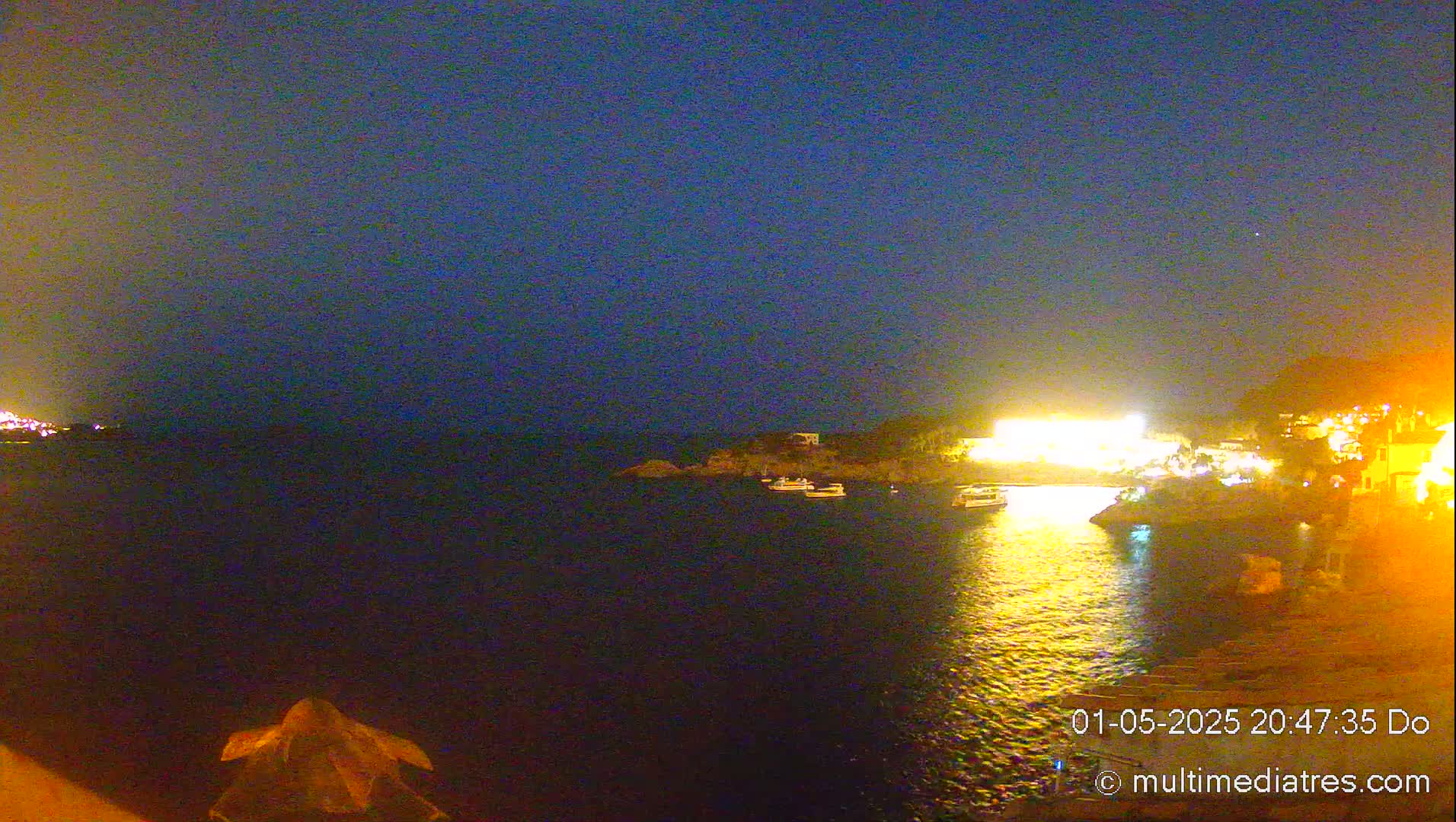 A nighttime view of a calm body of water with several boats moored near a brightly lit shoreline.