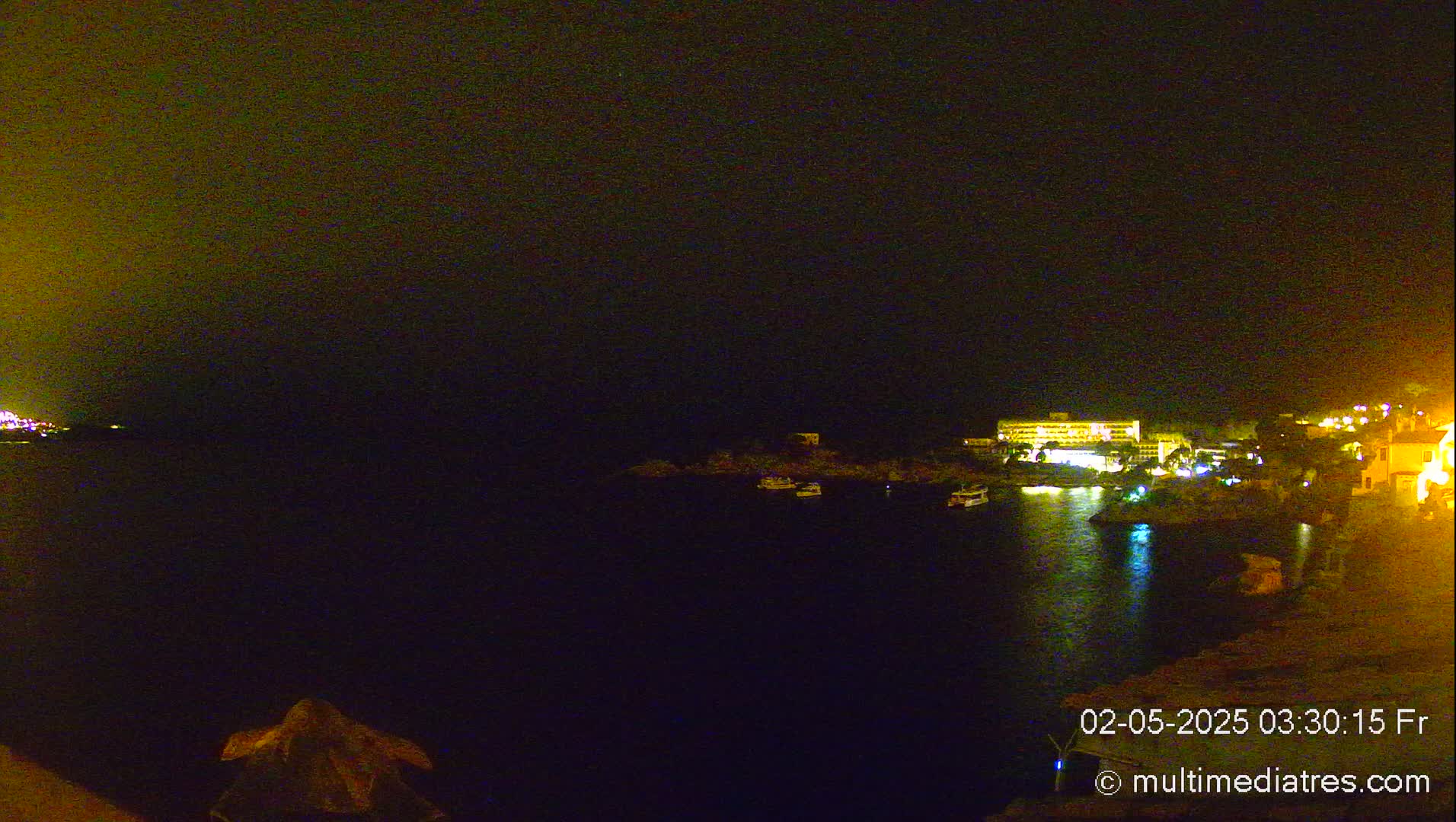 A nighttime view of a dark body of water with a few illuminated boats and a brightly lit building on the far shore under a dark sky.