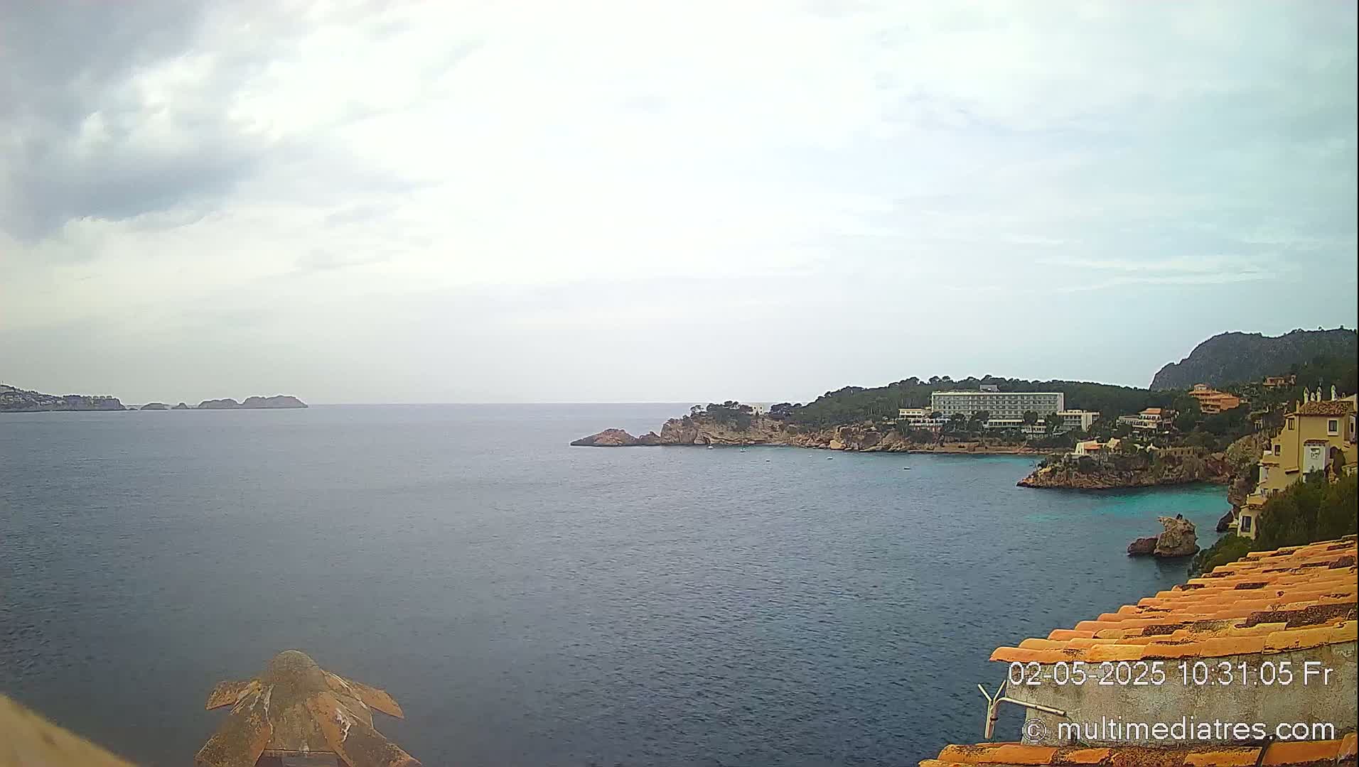 A coastal view shows a calm sea under an overcast sky, with a hotel and buildings situated on a rocky headland.