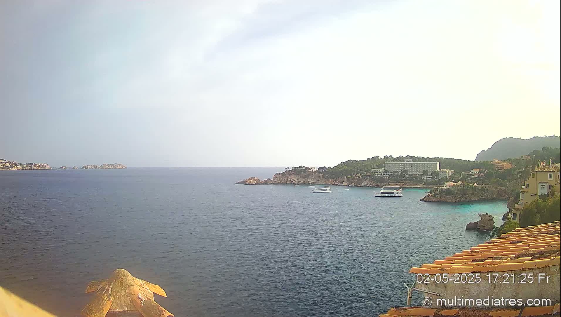 A calm, hazy sea stretches out to rocky islands and a coastline with buildings and trees under a light overcast sky.