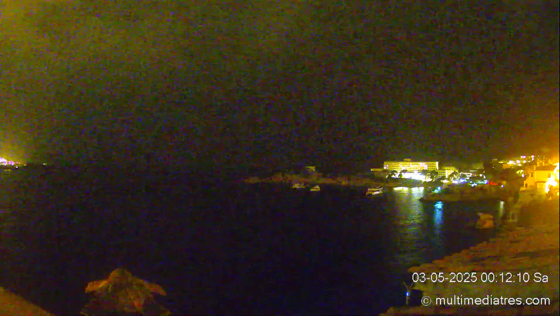 A nighttime view of a dark body of water with a few boats and illuminated buildings on the shore under a dark, possibly cloudy sky.