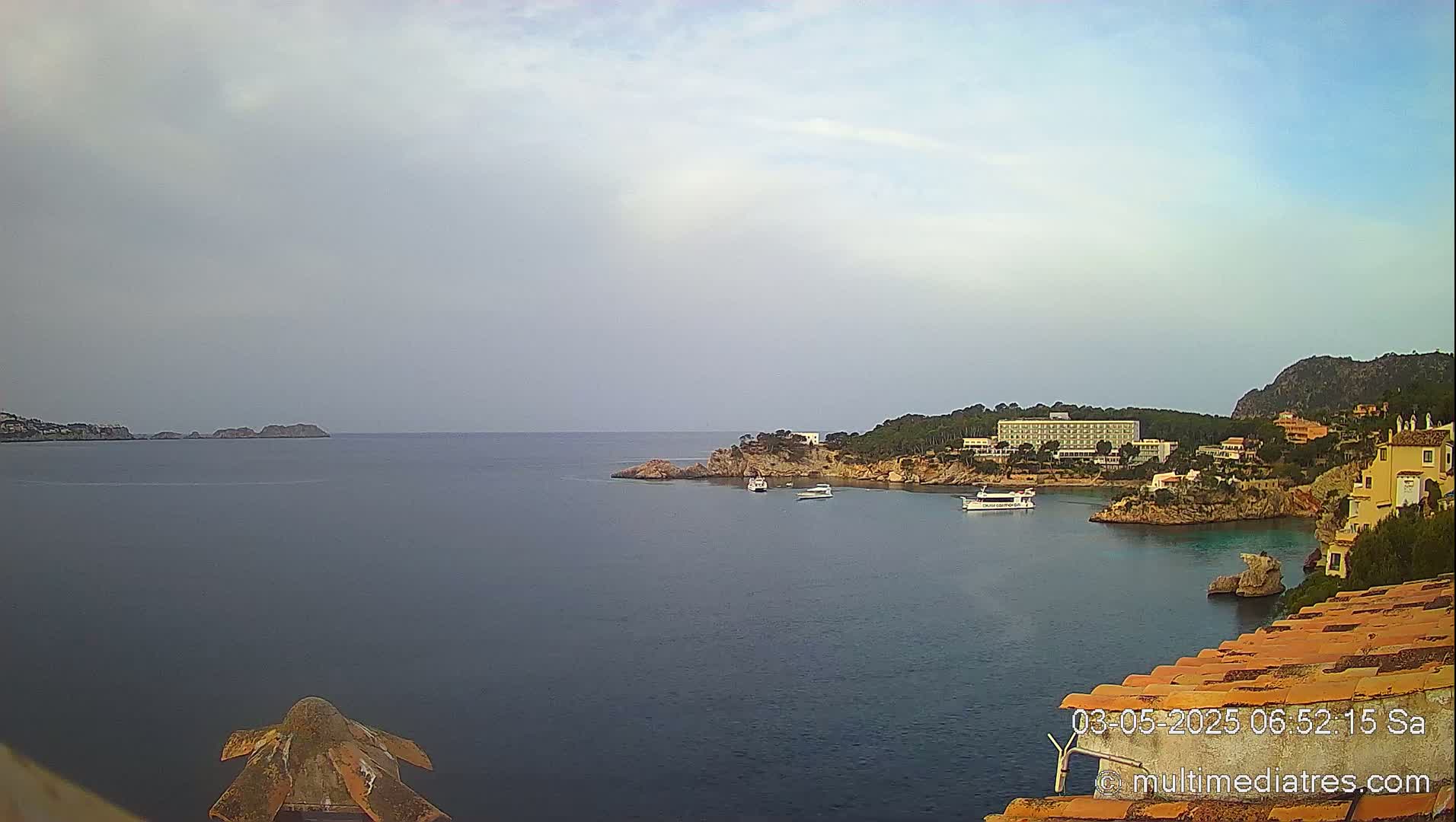 A calm, overcast day reveals a coastal scene with a large hotel and several buildings nestled on a rocky peninsula jutting into a bay, with boats visible in the water and smaller islands in the distance.