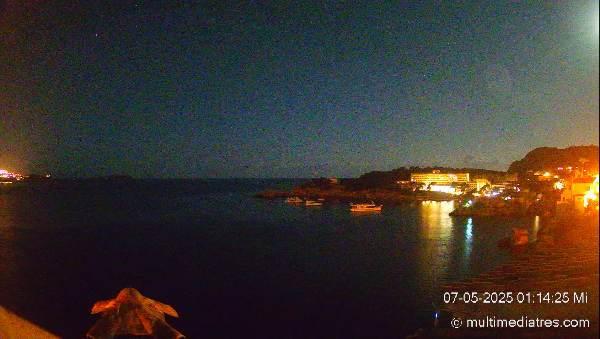 A nighttime view of a calm bay with a few boats, illuminated buildings on the shore, and a starry sky.