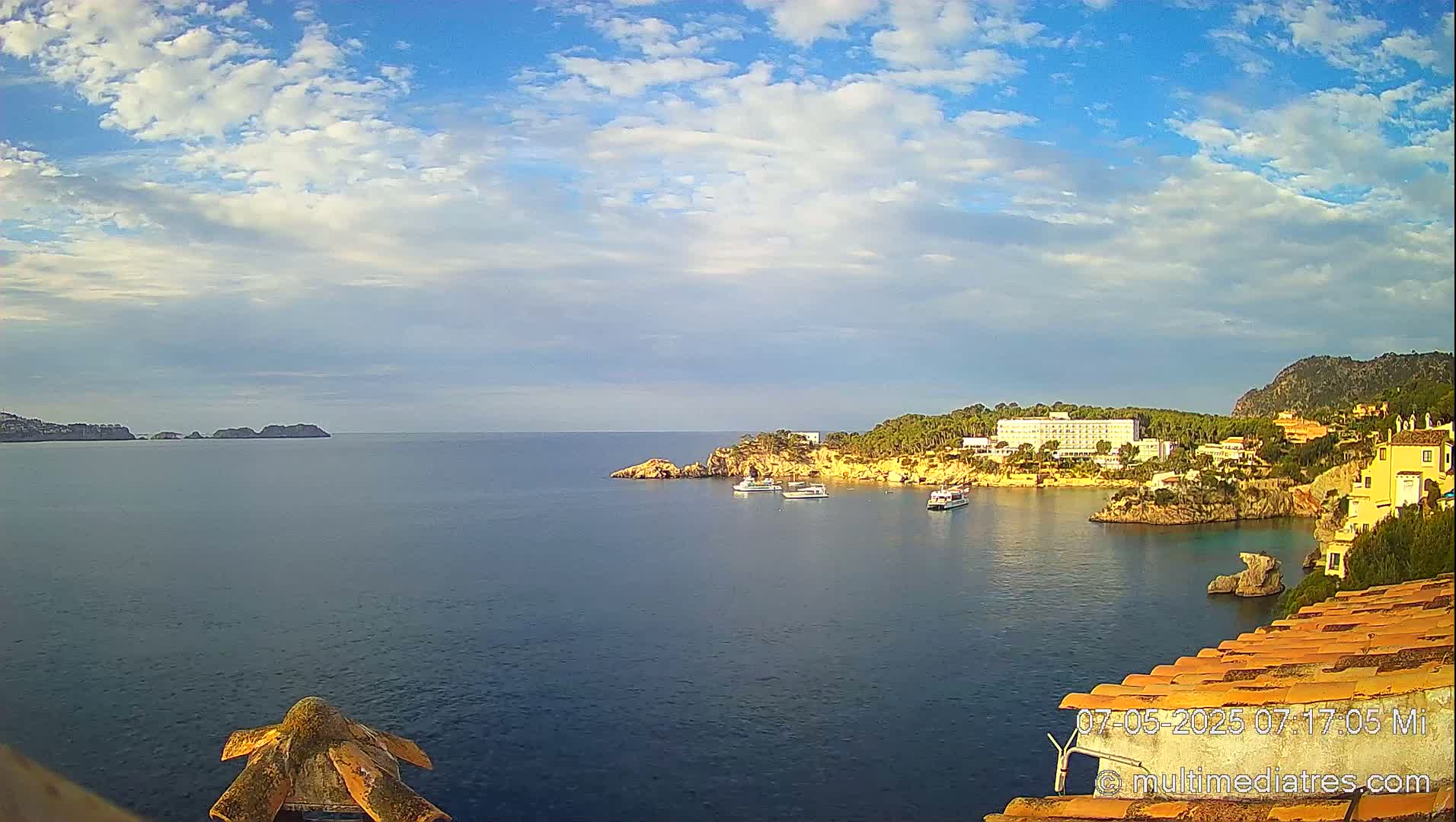 A calm bay with several boats, a large hotel, and other buildings is seen under a partly cloudy sky.