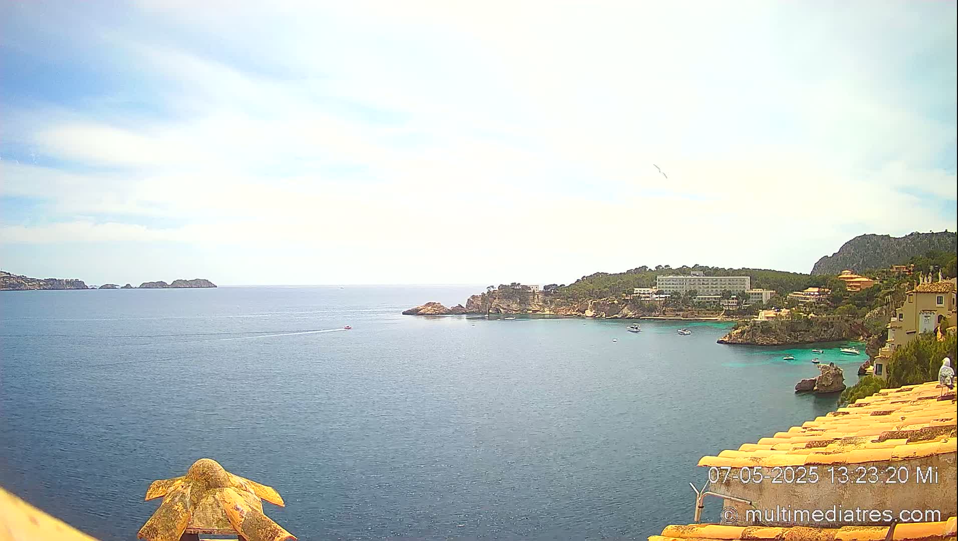 A partly cloudy sunny day overlooks a calm ocean bay with a rocky coastline featuring buildings and vegetation.