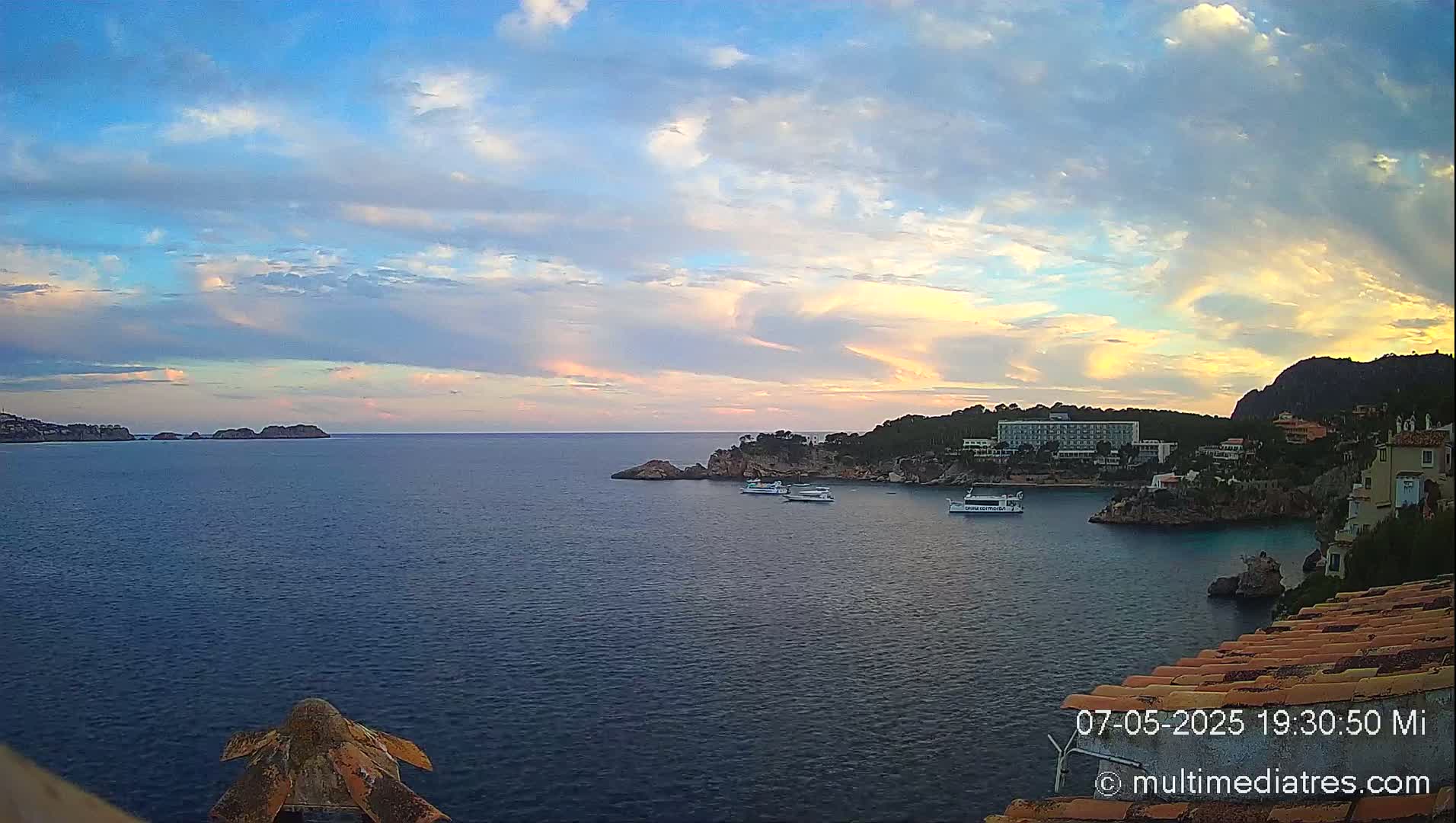 A coastal scene at sunset shows a calm sea with several boats, rocky coastlines, and buildings nestled into the hills under a partly cloudy sky.