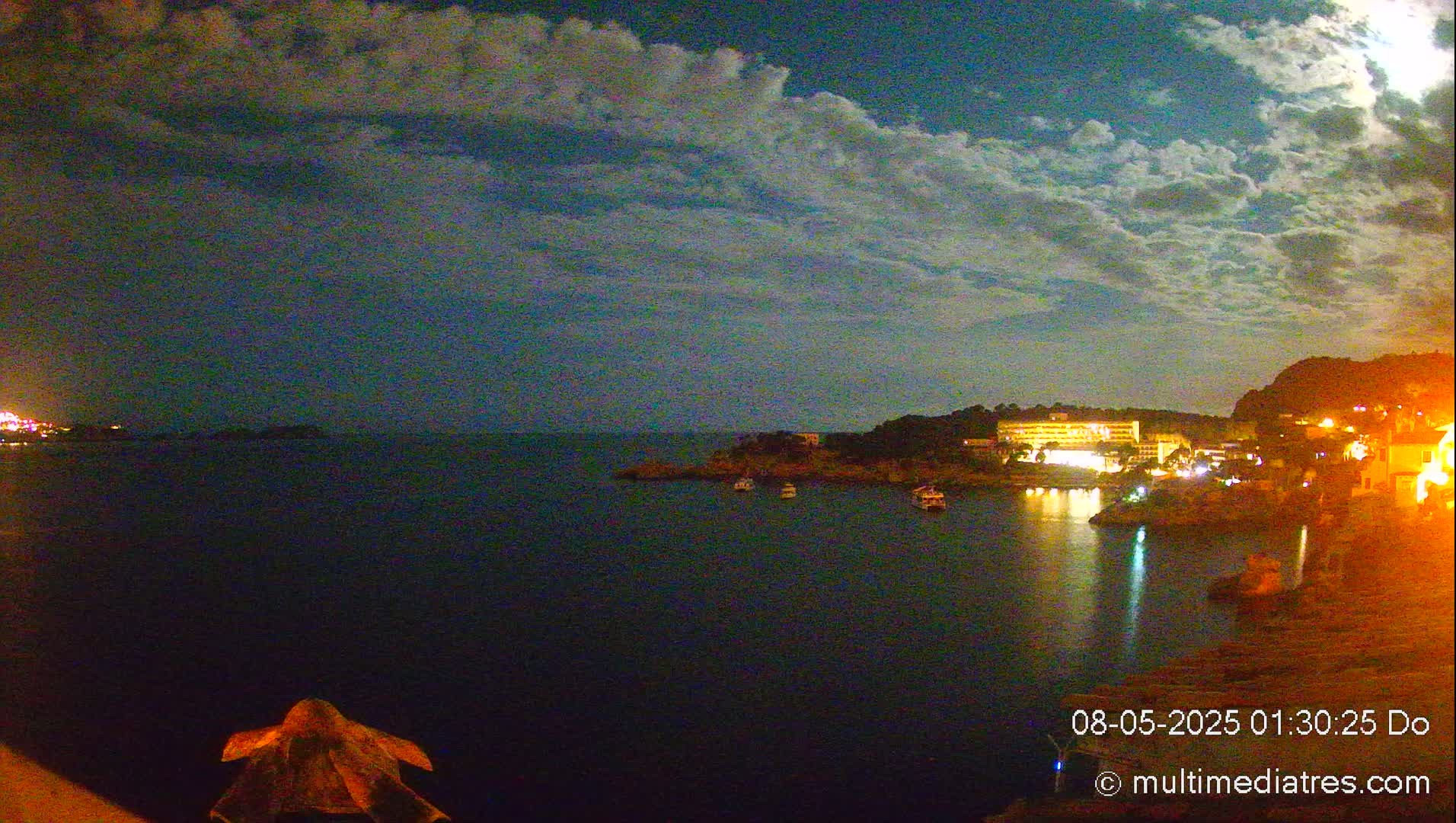 A nighttime coastal scene shows a calm body of water with a few boats, illuminated buildings on the shore, and a partly cloudy sky.
