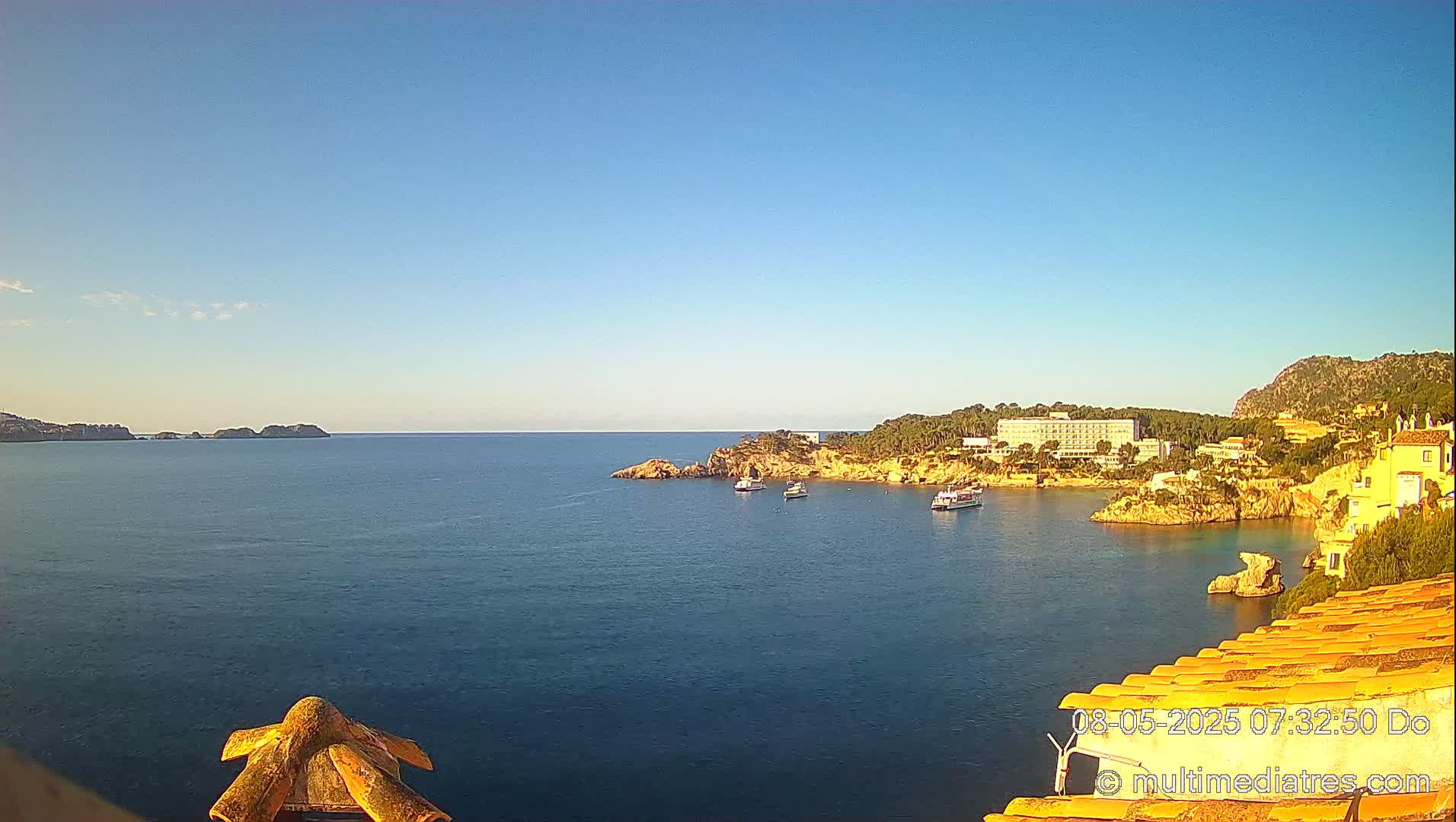 A calm, blue sea stretches out to distant islands under a clear, sunny sky, with several boats in a small bay near a hotel and buildings on a rocky coastline.